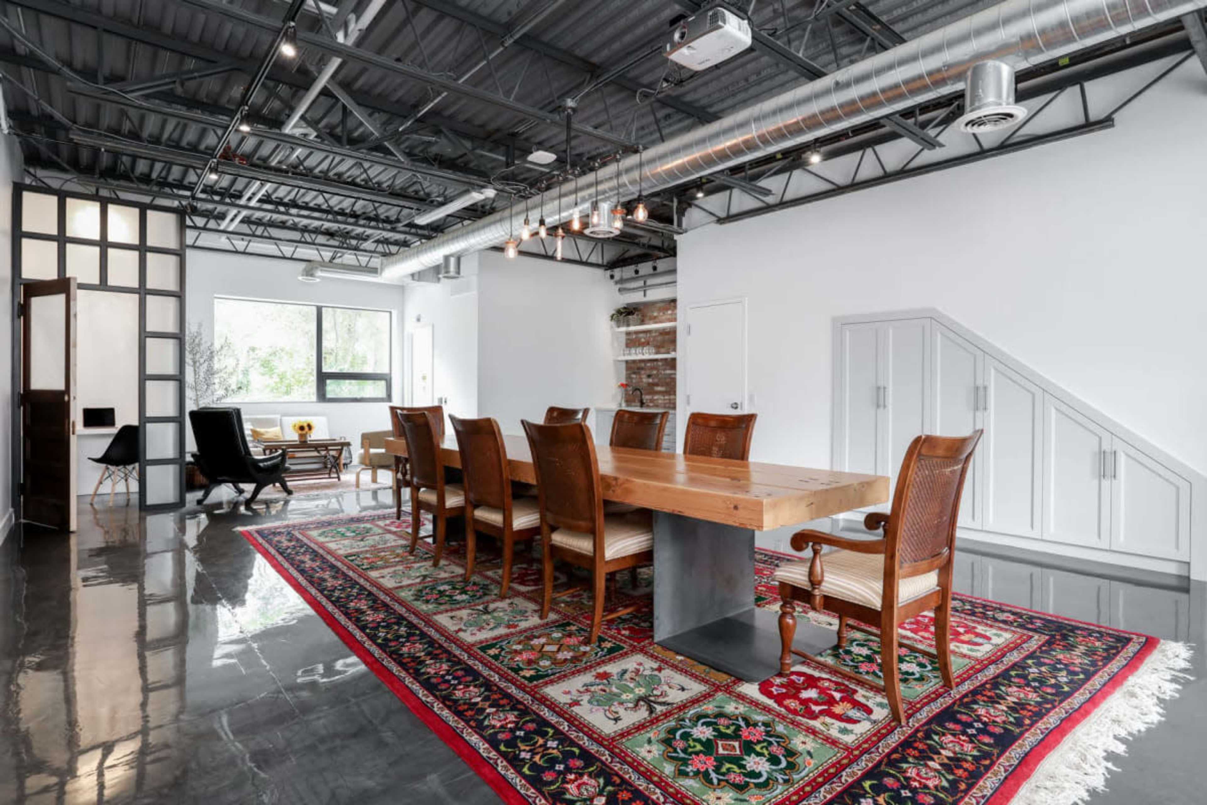 A modern dining area with a large wooden table surrounded by eight chairs, set on a colorful area rug, within a room featuring industrial-style decor and natural light.
