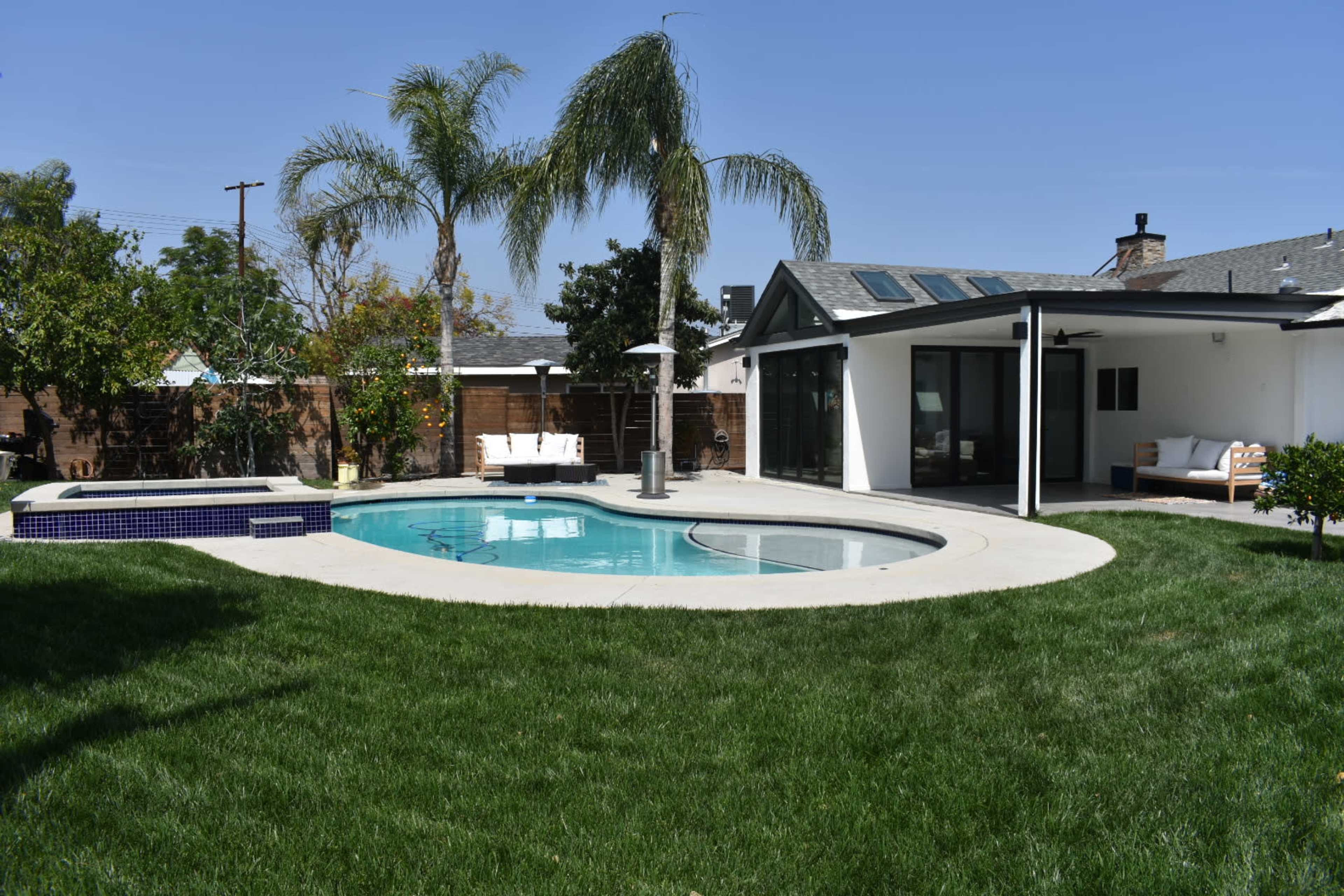 A backyard with a swimming pool, a hot tub, and a modern house surrounded by green grass and palm trees.