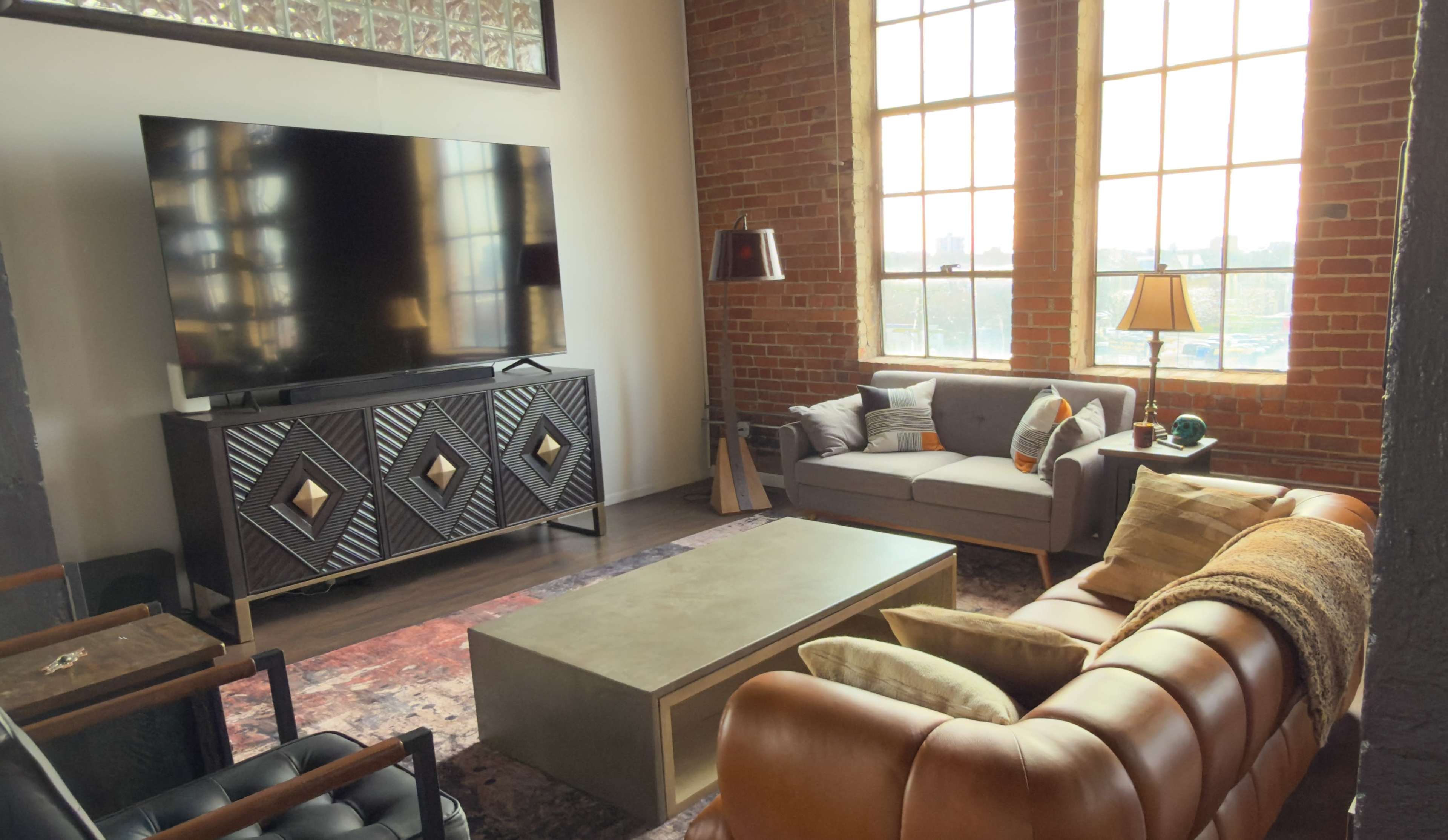 The image shows a modern living room with large windows, a flat-screen TV on a patterned cabinet, a light-colored coffee table, and a mix of seating options including a brown leather couch and a gray sofa.