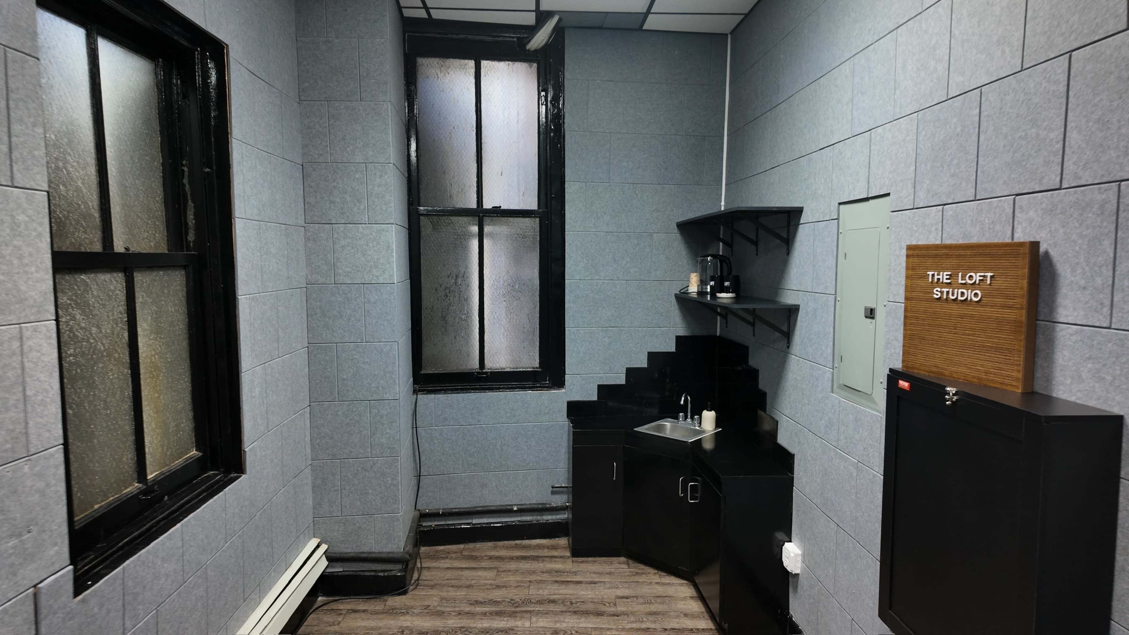 The image shows a small room with gray walls, a kitchenette area featuring a sink, a black cabinet, and a wooden sign that reads "THE LOFT STUDIO."
