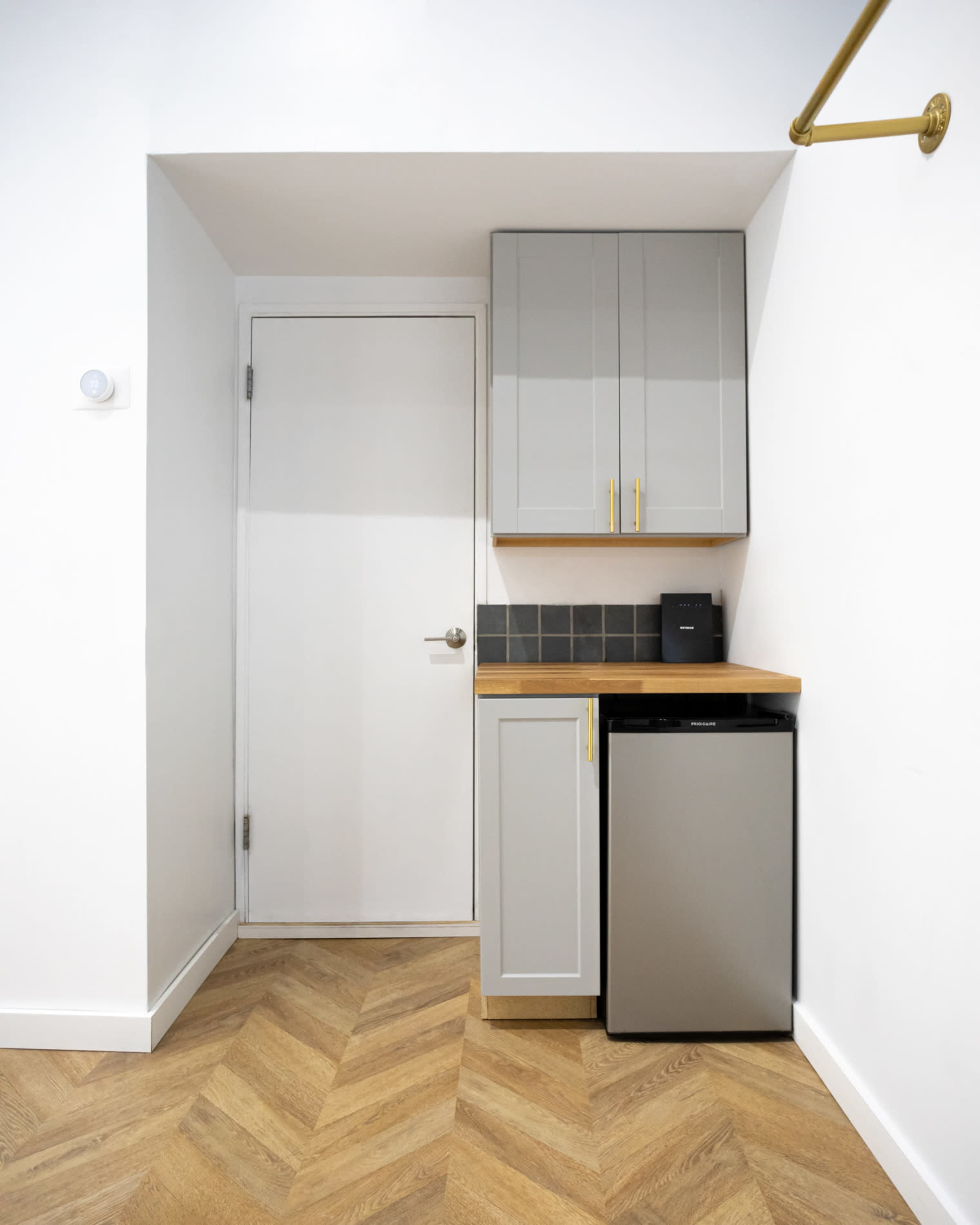 The image shows a small kitchen area featuring grey cabinets, a wooden countertop, a mini fridge, and a white door against a herringbone floor pattern.