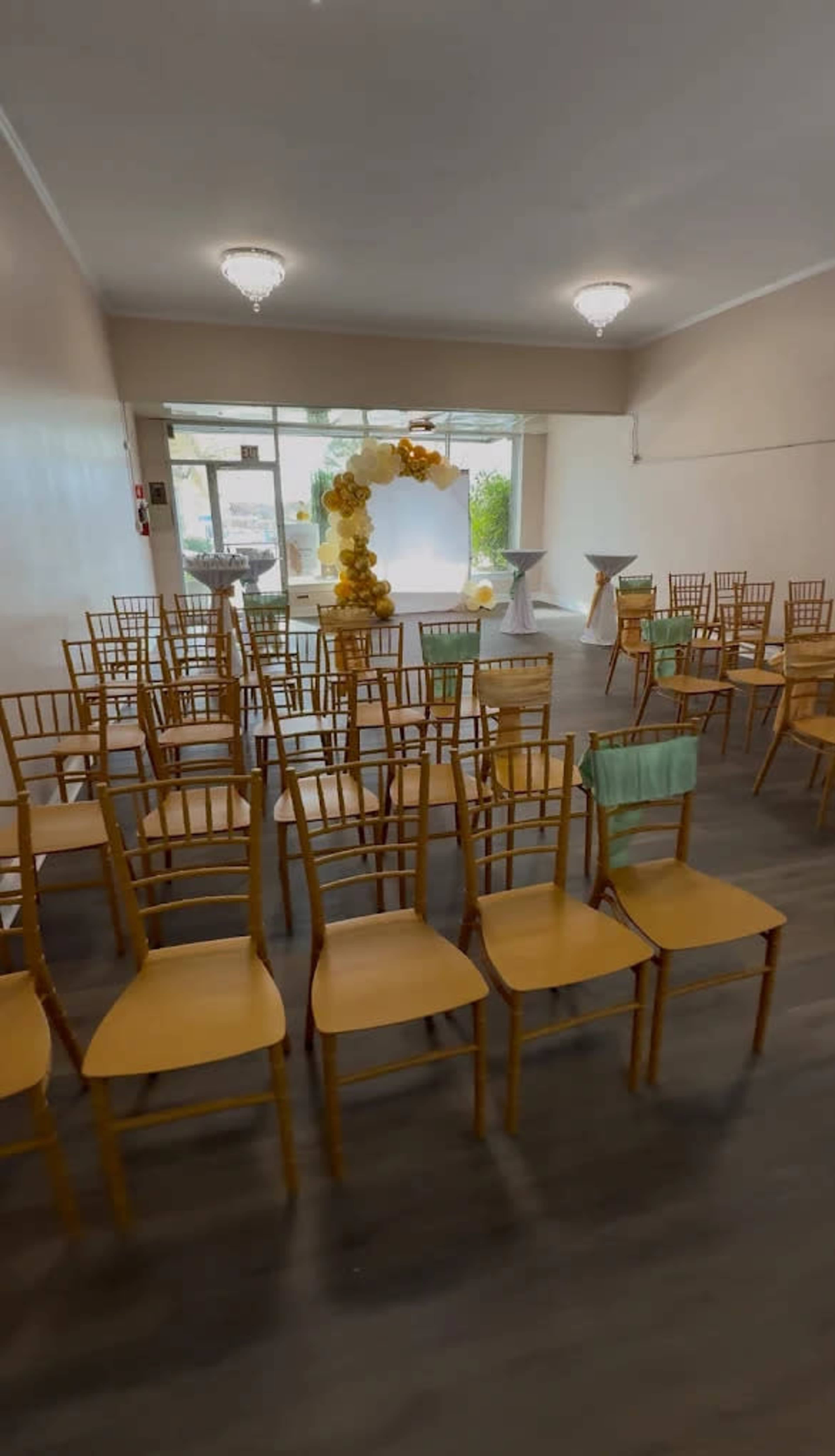 The image shows a spacious room set up for an event, featuring rows of wooden chairs arranged facing a decorated backdrop with a table and floral arrangements in the background.