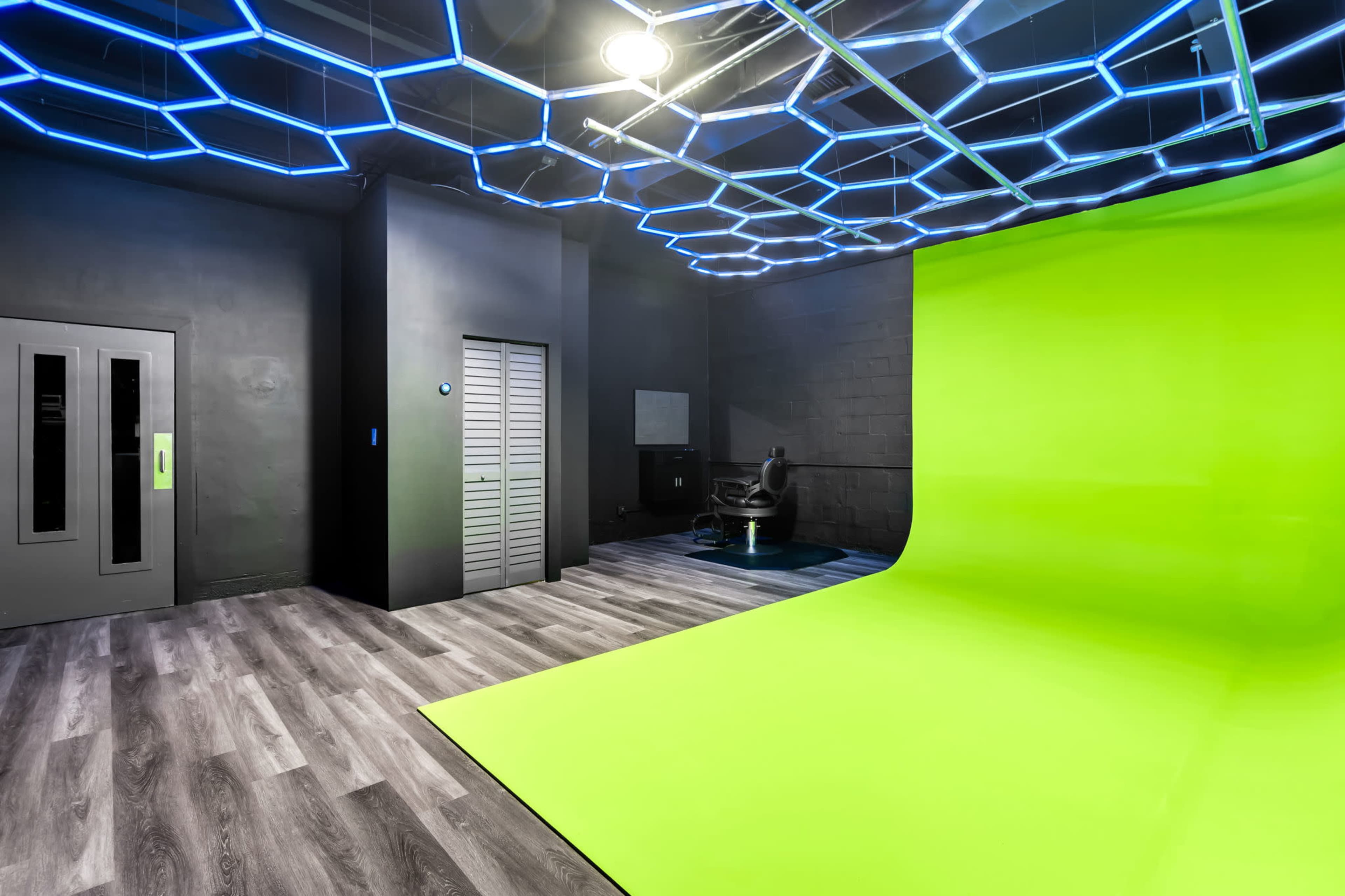The image shows a studio with a bright green backdrop, dark walls, a stylish ceiling with blue hexagonal lights, and a black chair positioned in the corner.
