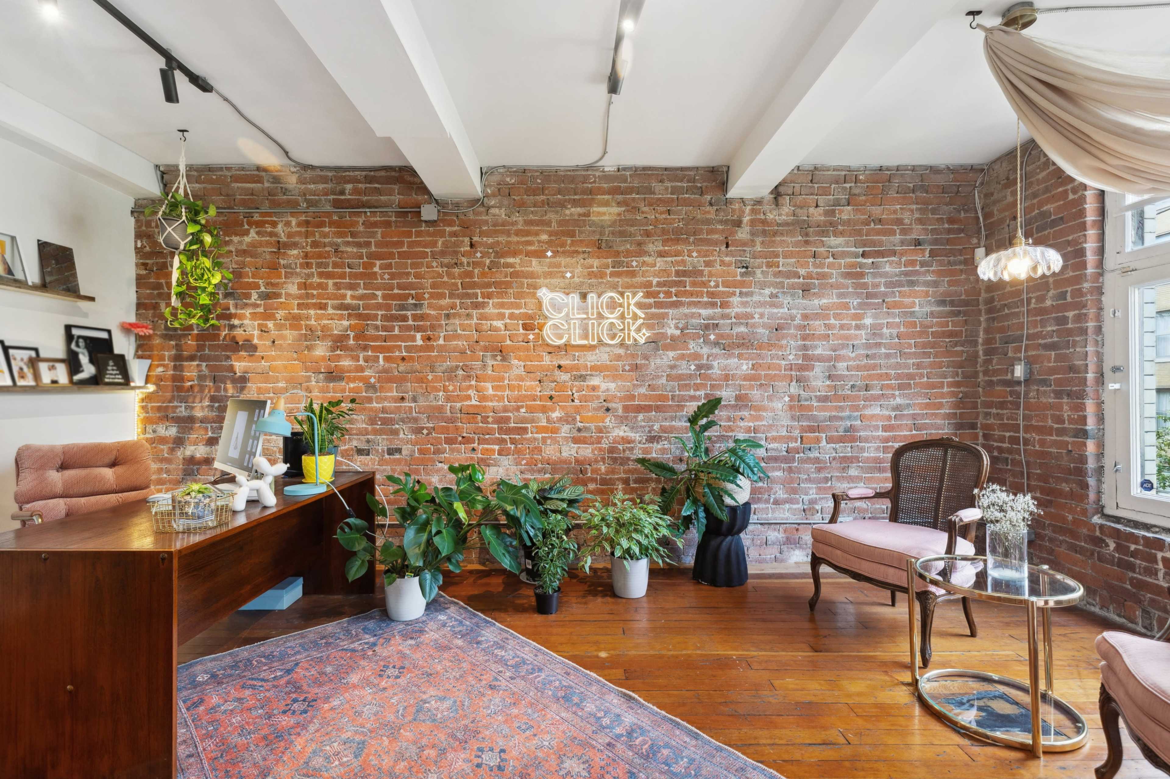 A modern office space with exposed brick walls, a wooden desk, and various potted plants, featuring a decorative rug and seating arrangements.