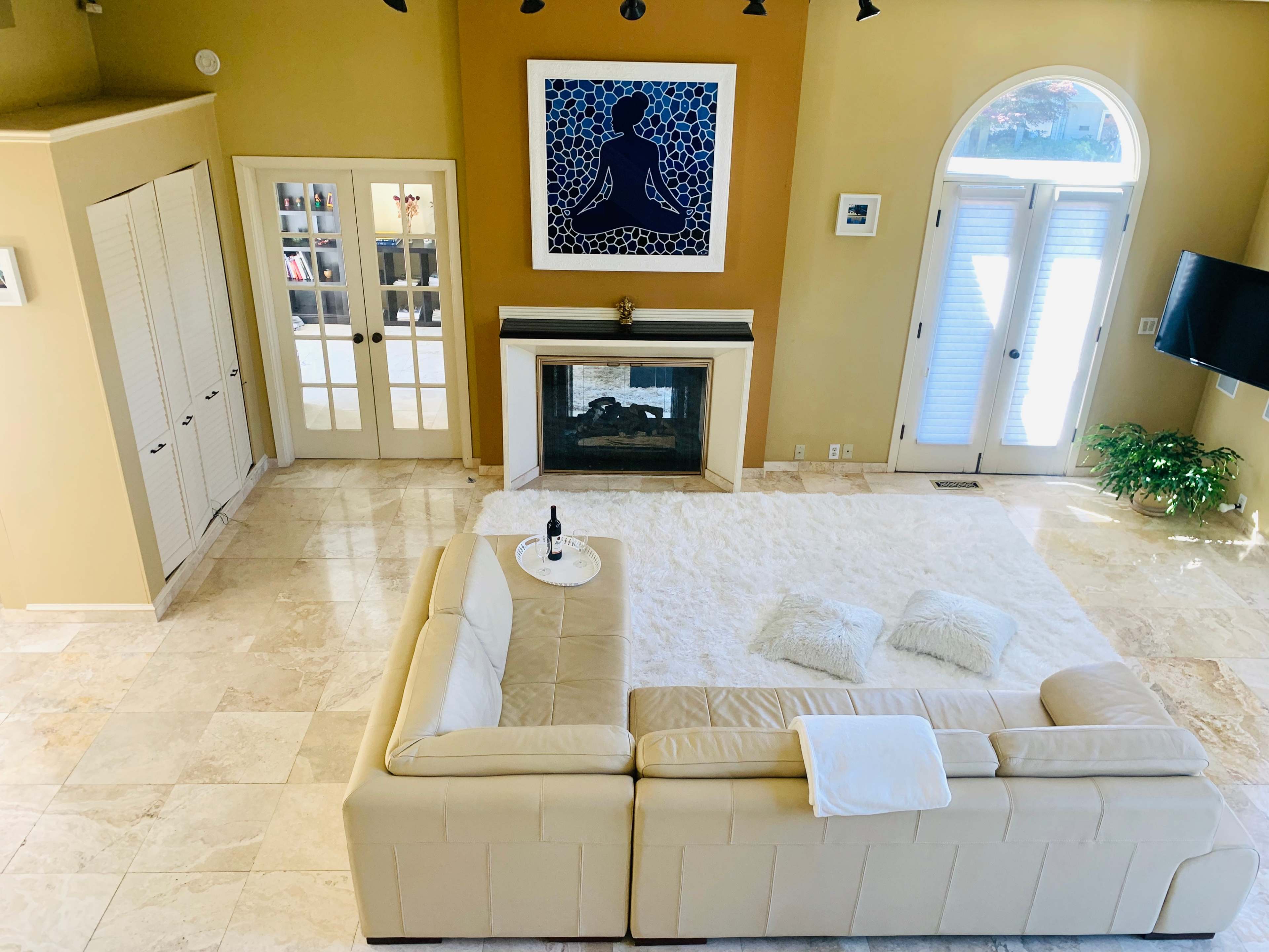 The room features a large beige sectional sofa, a white area rug, a fireplace, and a framed artwork on the wall, with double doors leading to another room.