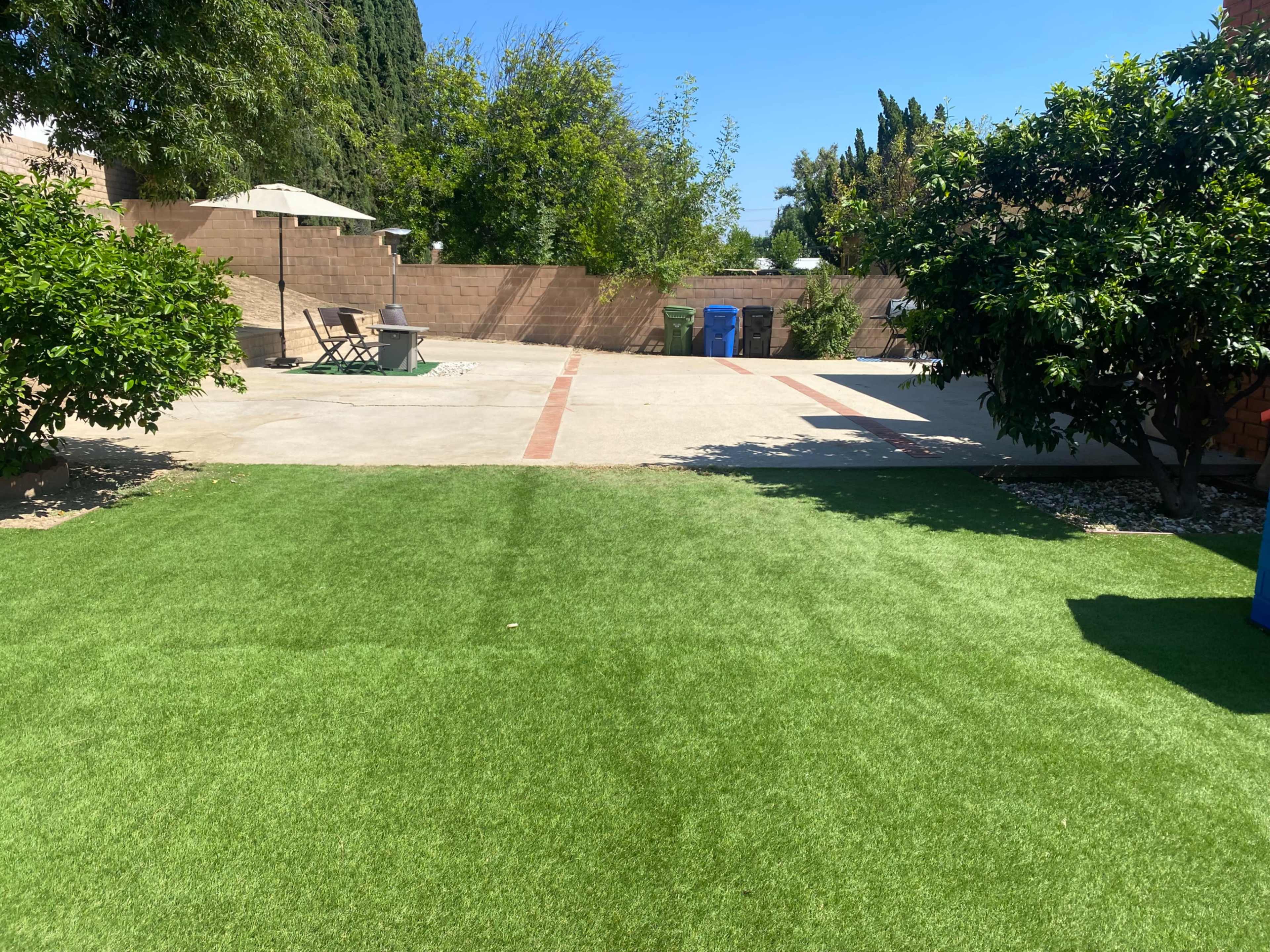 The image shows a backyard with artificial turf in the foreground, a paved area in the background, and a patio with an umbrella and seating.