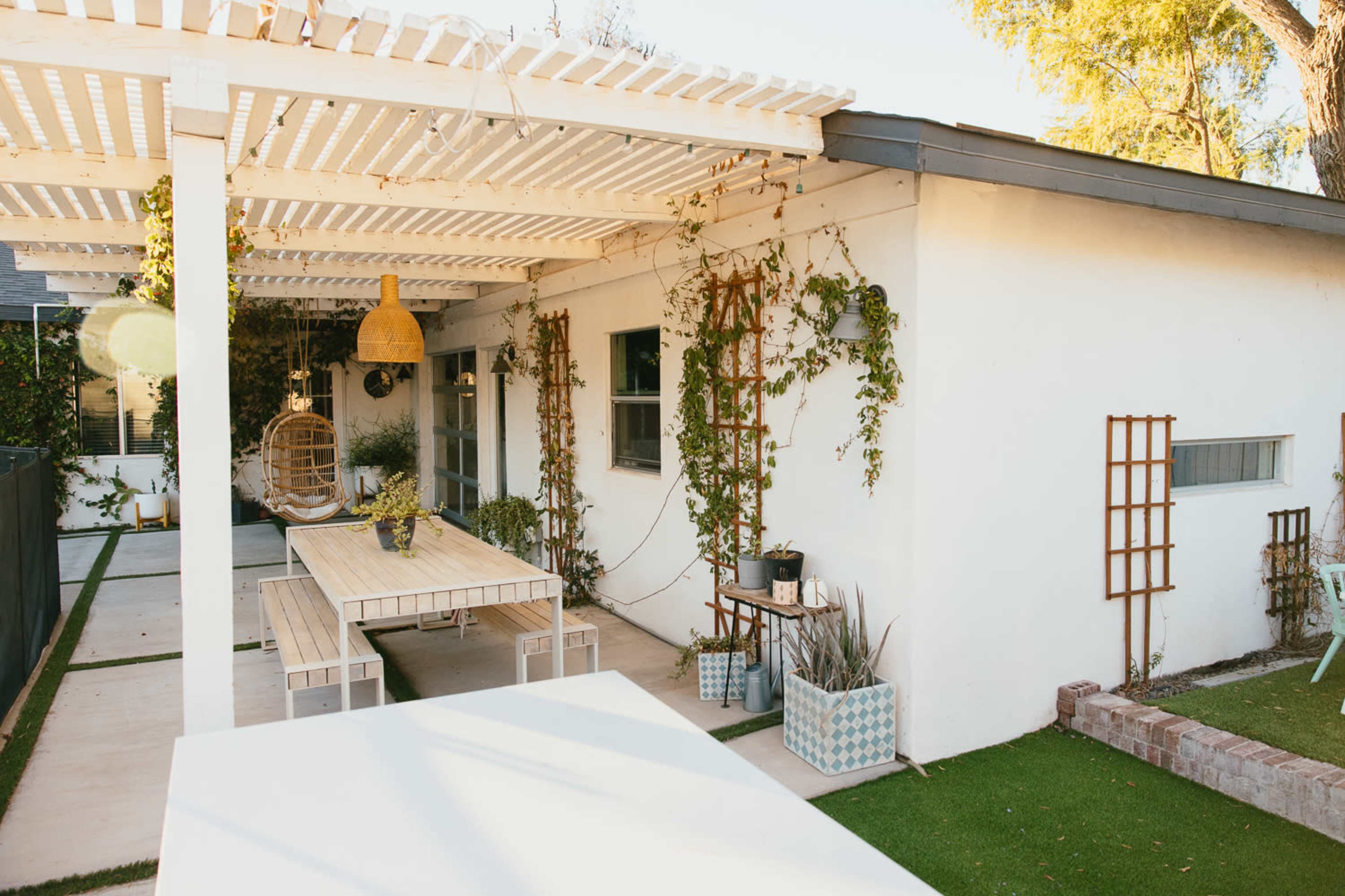 A patio with a white, slatted pergola, a long table surrounded by chairs, and walls adorned with climbing plants.