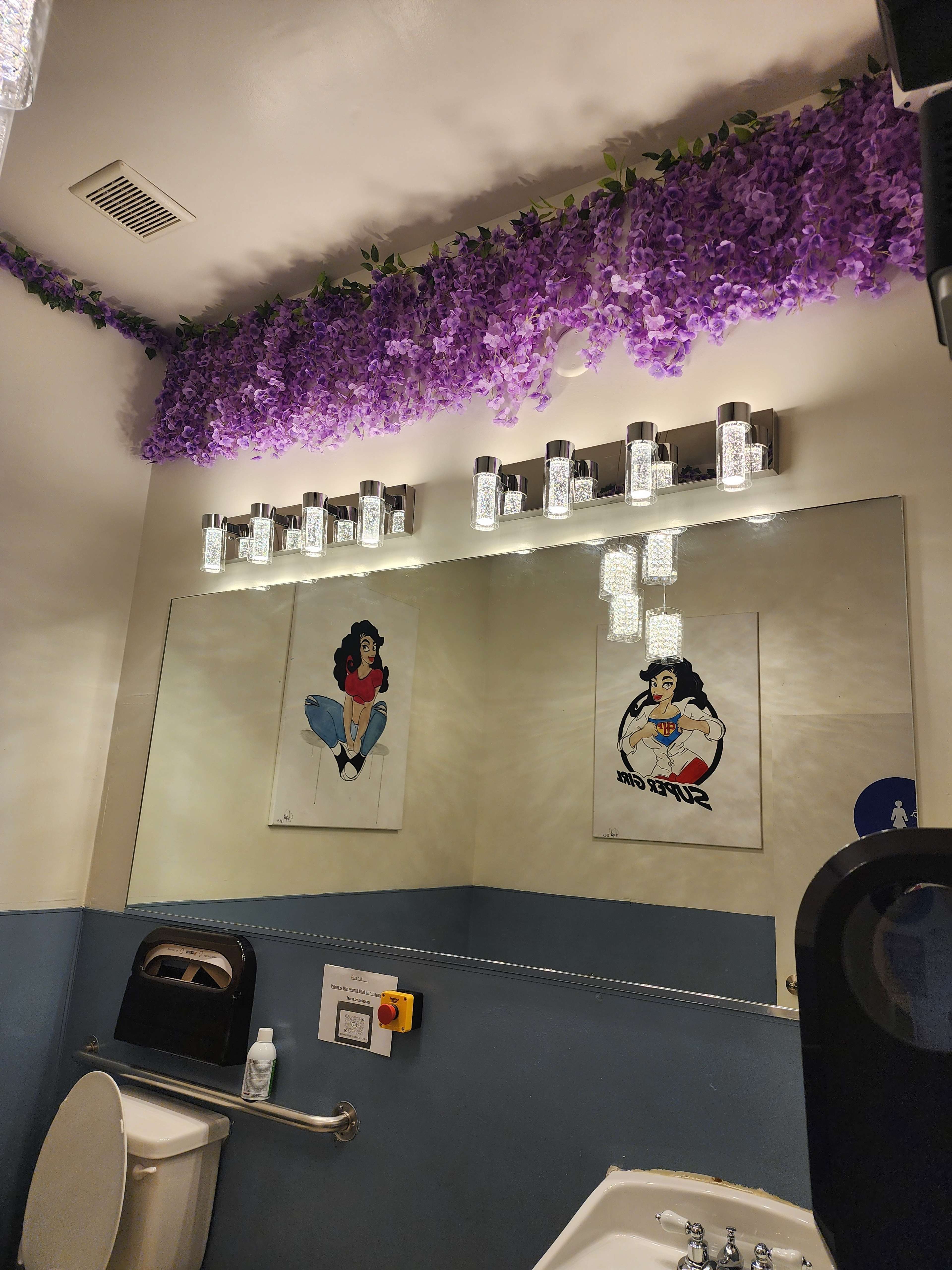 The image shows a bathroom with purple floral decorations along the ceiling, two illustrated posters on the wall, a light fixture above the mirror, and standard bathroom fixtures.
