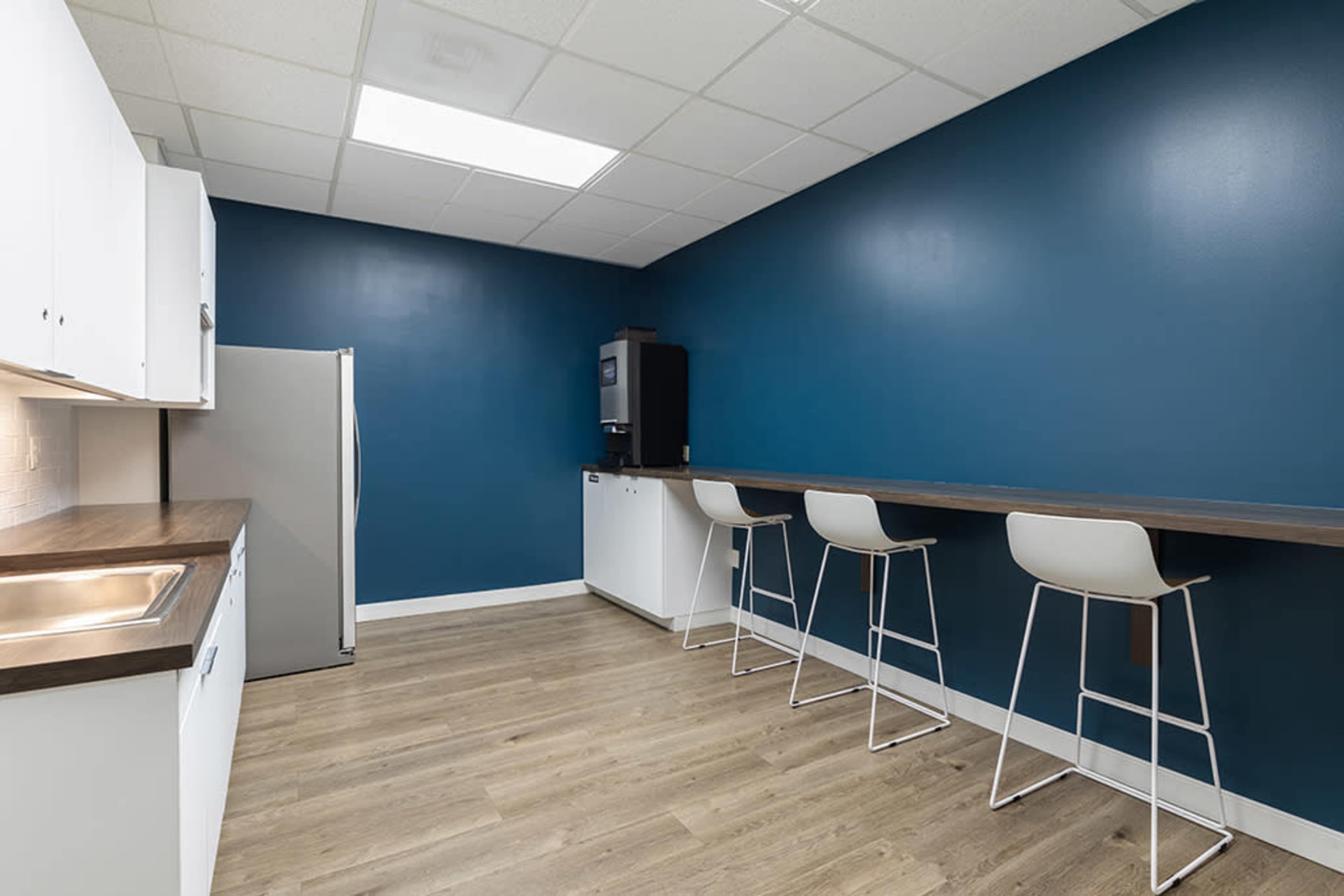 The image shows a modern kitchen with a blue accent wall, white cabinets, a silver refrigerator, and a bar area with three white stools.