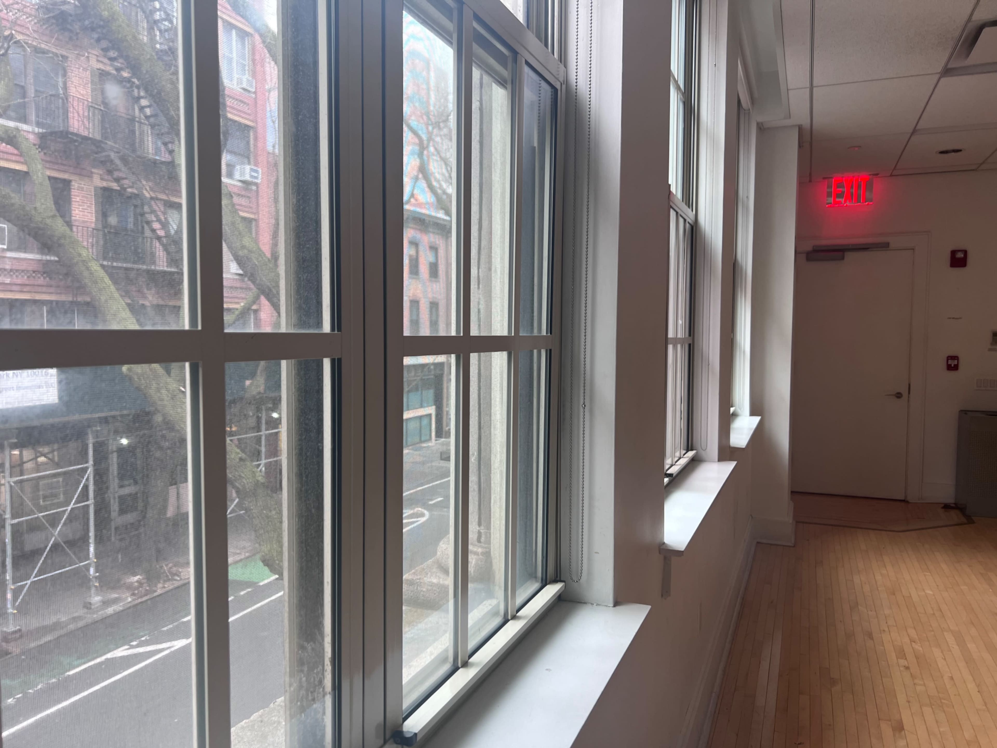 The image shows a bright interior space with large windows alongside a wooden floor, revealing a view of a street and buildings outside.
