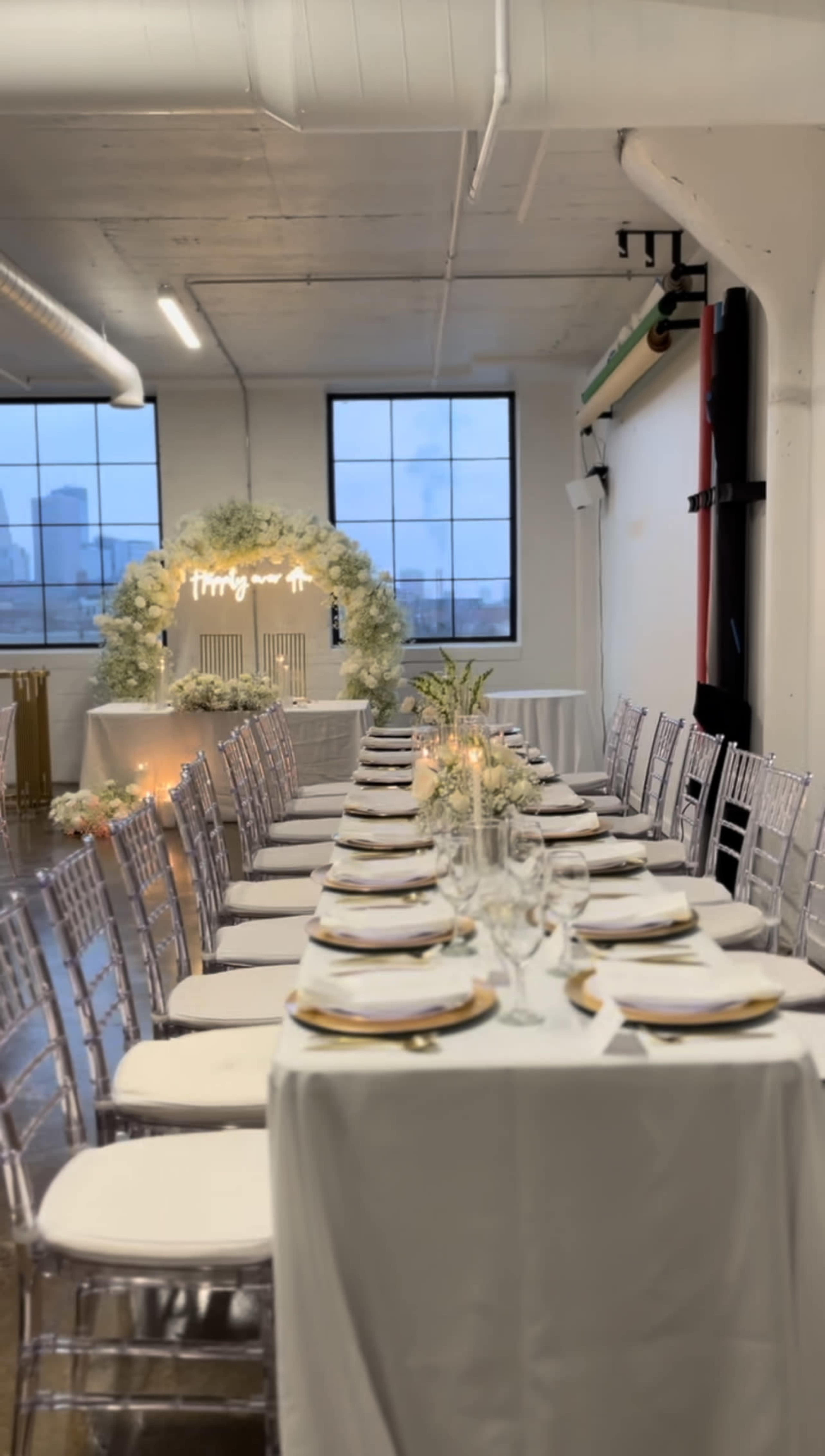 A long dining table is set with plates and glasses in a decorated event space featuring large windows and floral arrangements.