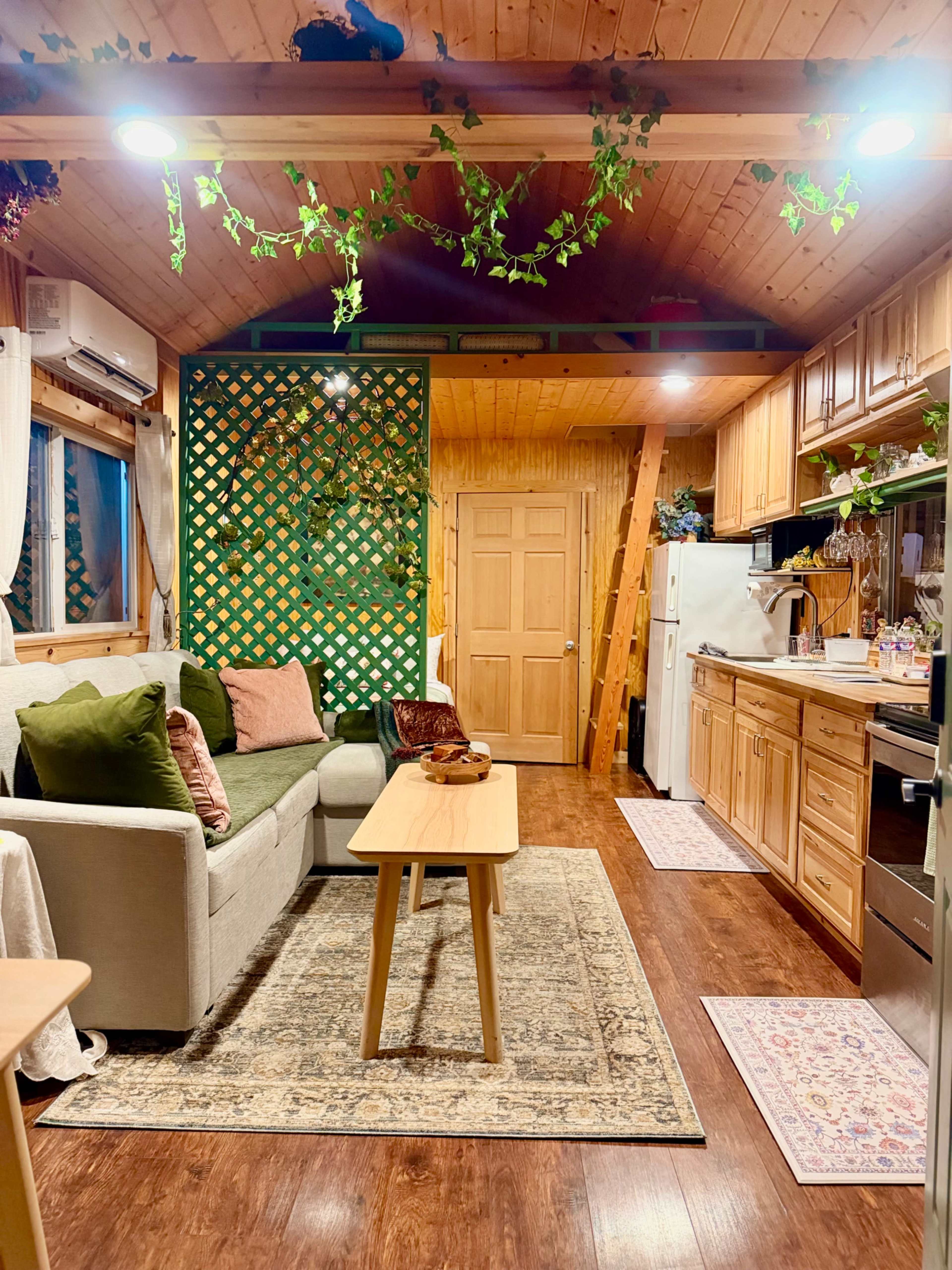 The image shows a cozy living space with a sofa, a wooden table, and a kitchen area adorned with green plants and wooden cabinetry.