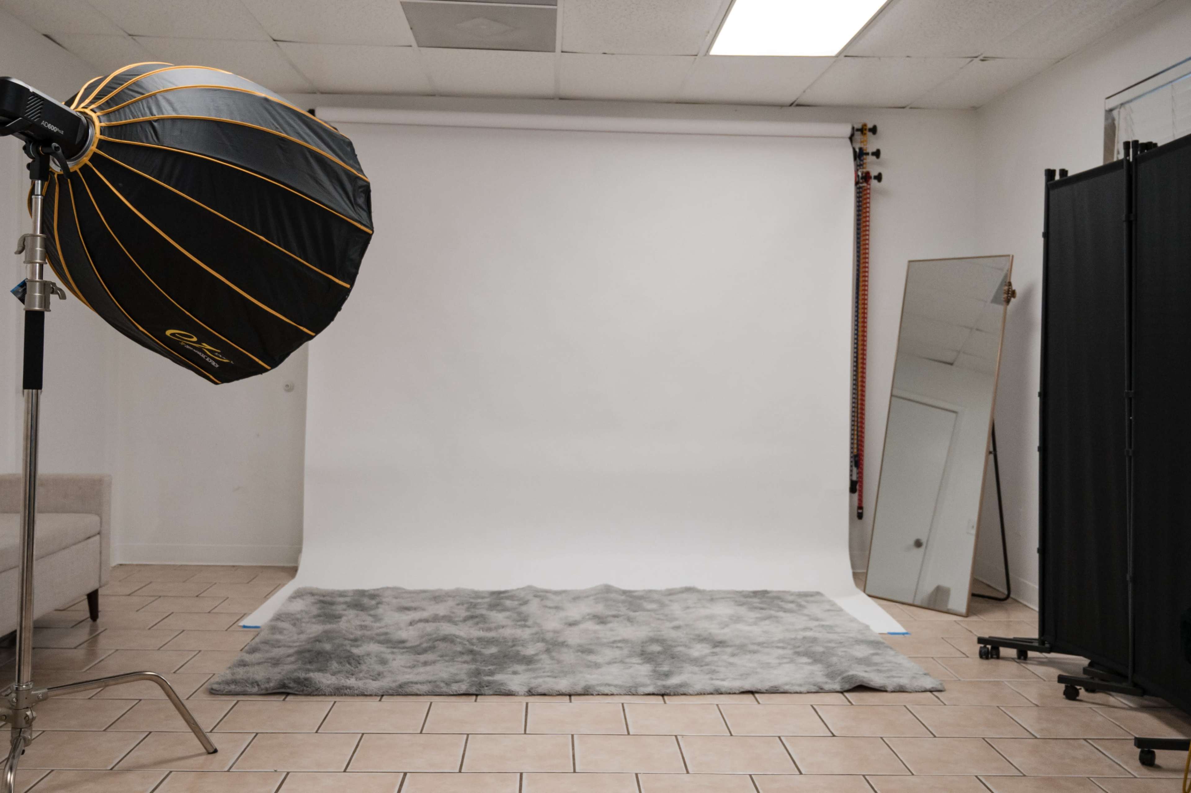 A photography studio features a softbox light, a large mirror, and a gray rug placed on tiled flooring against a plain white backdrop.