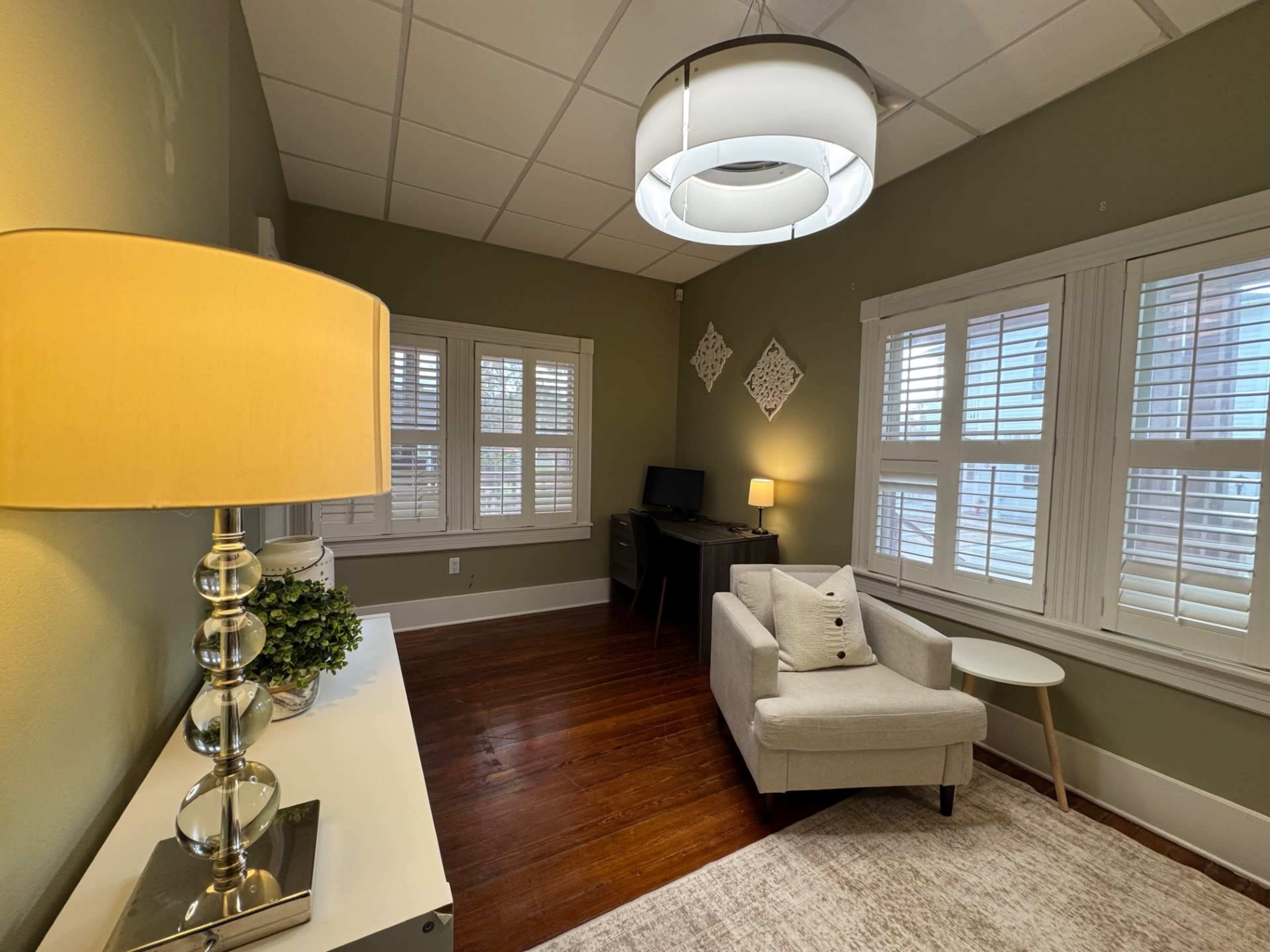 The image shows a small, well-lit room with a plush chair, a desk, and large windows with shutters, featuring a lamp and decorative items.