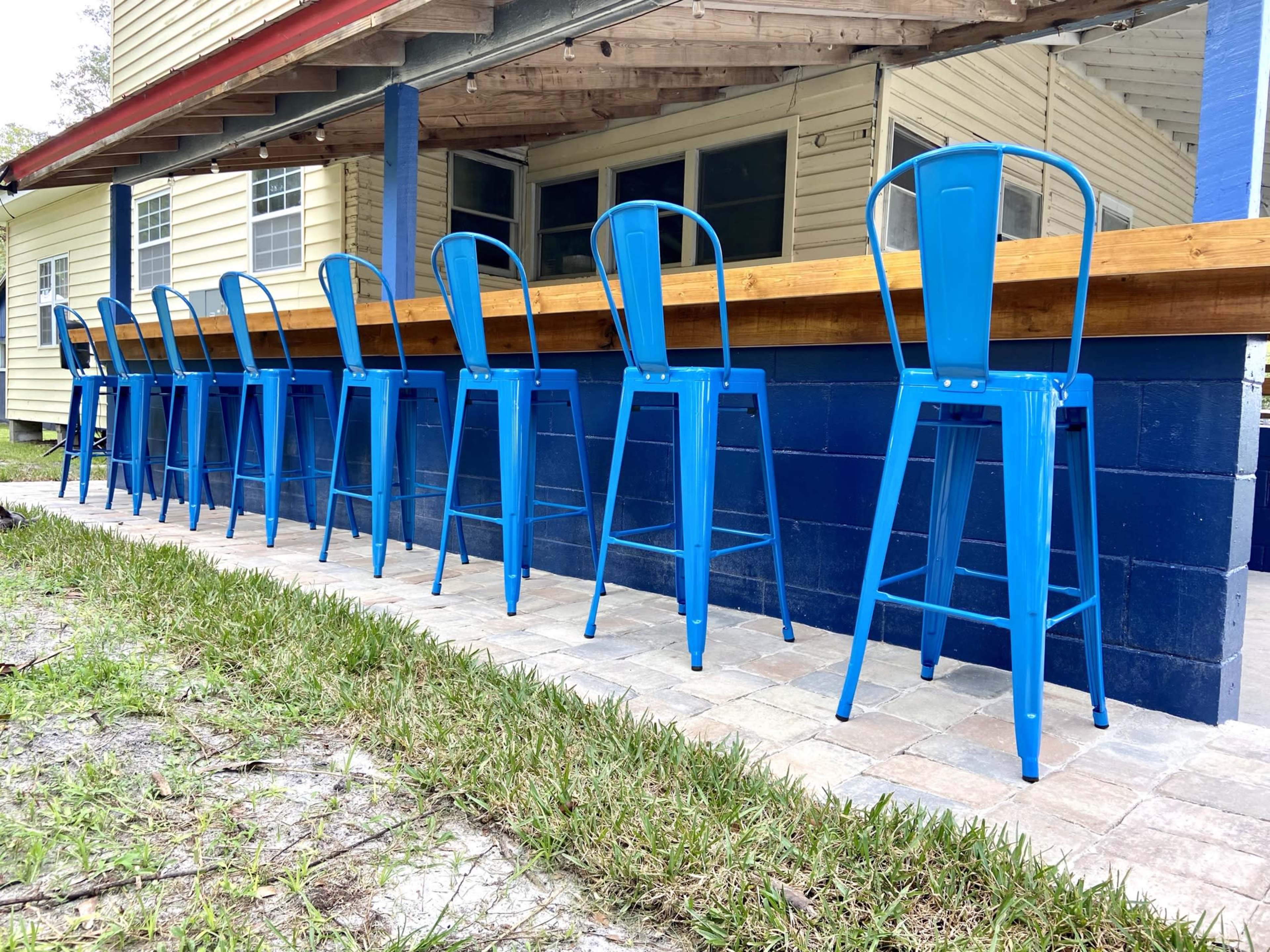 A row of blue metal bar stools is aligned along a wooden counter outside a building with yellow siding.