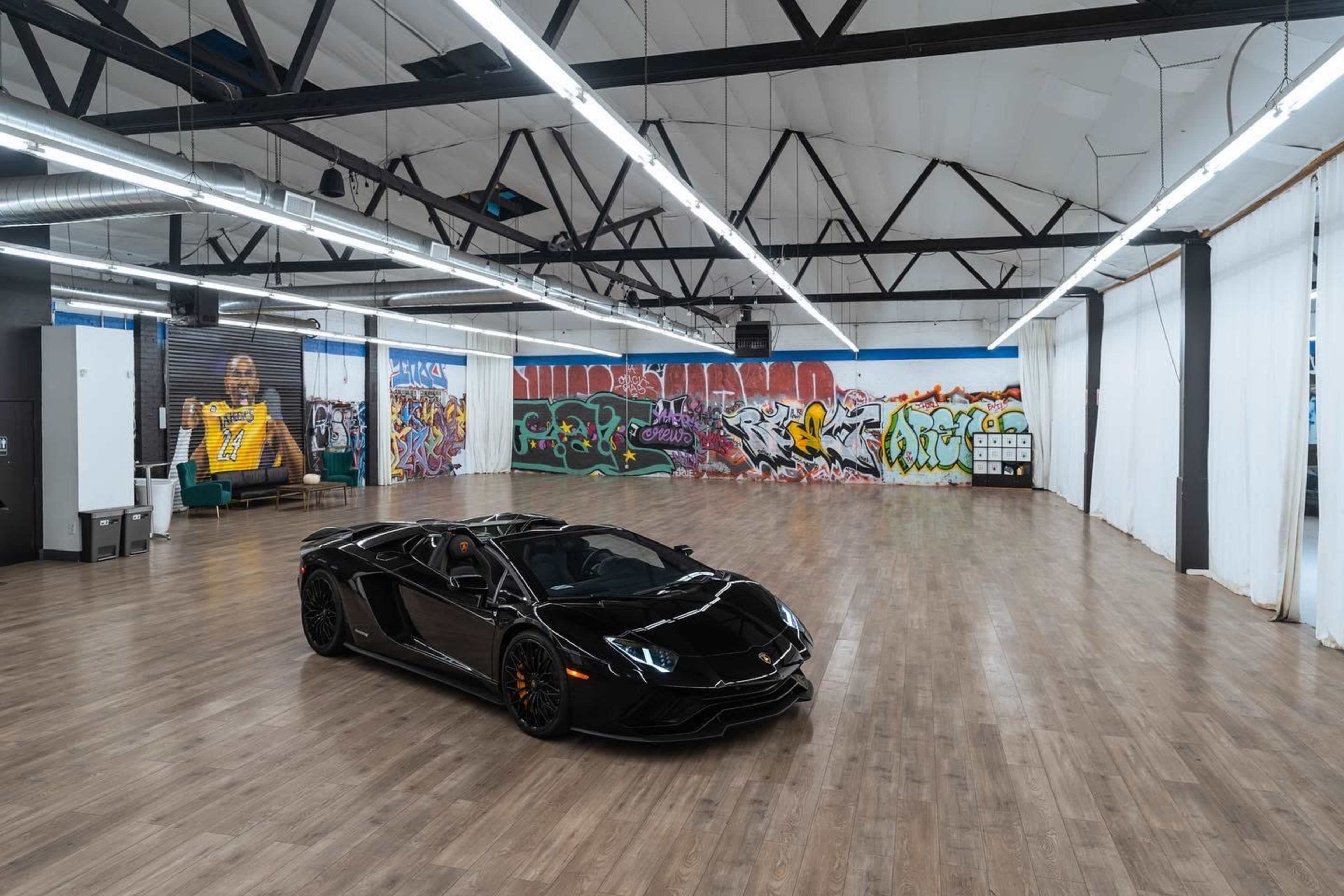 A black sports car is parked in an empty industrial-style room with wooden floors and colorful graffiti on the walls.