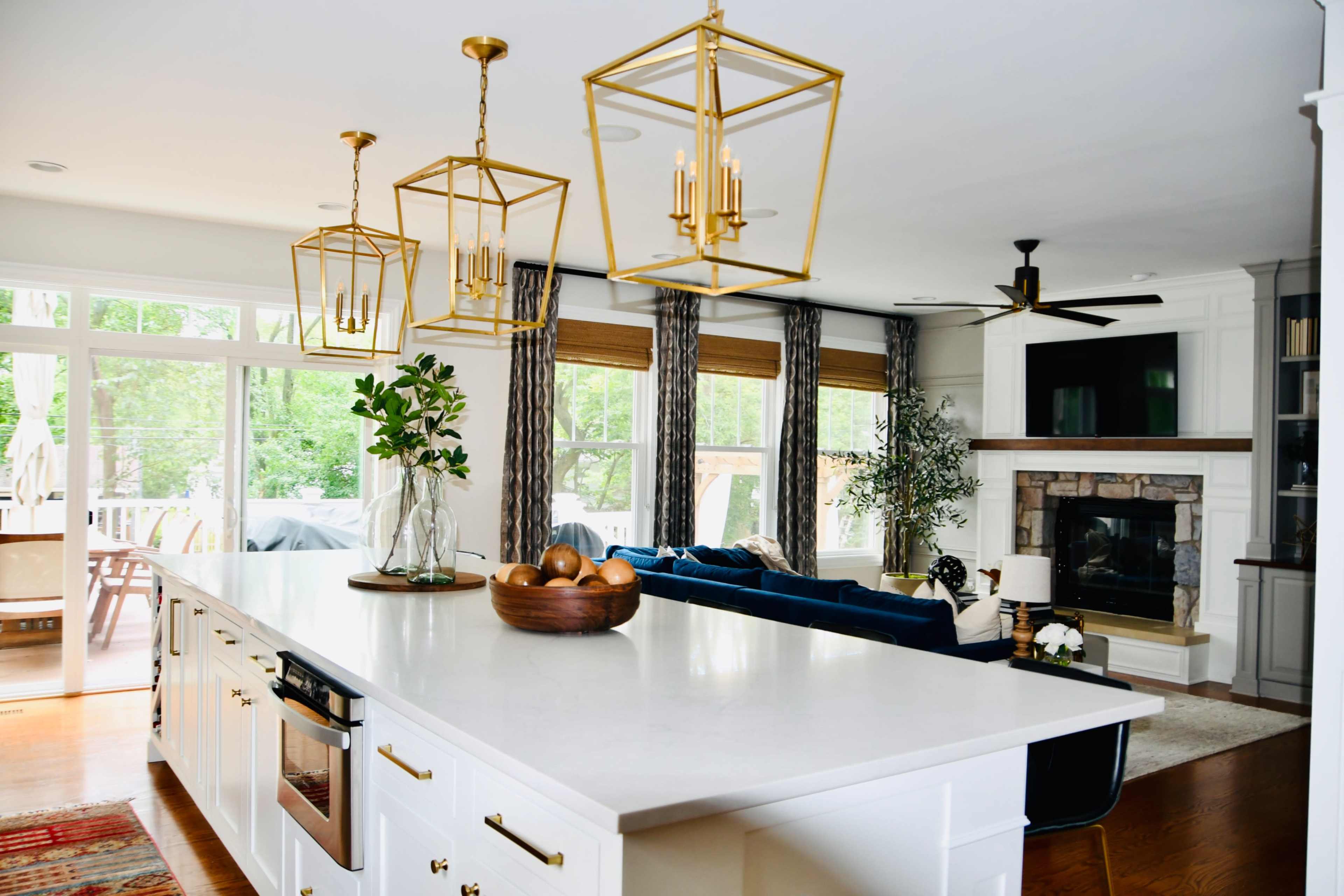 A modern kitchen with a large white island, brass pendant lights, and a living area featuring a fireplace and seating.