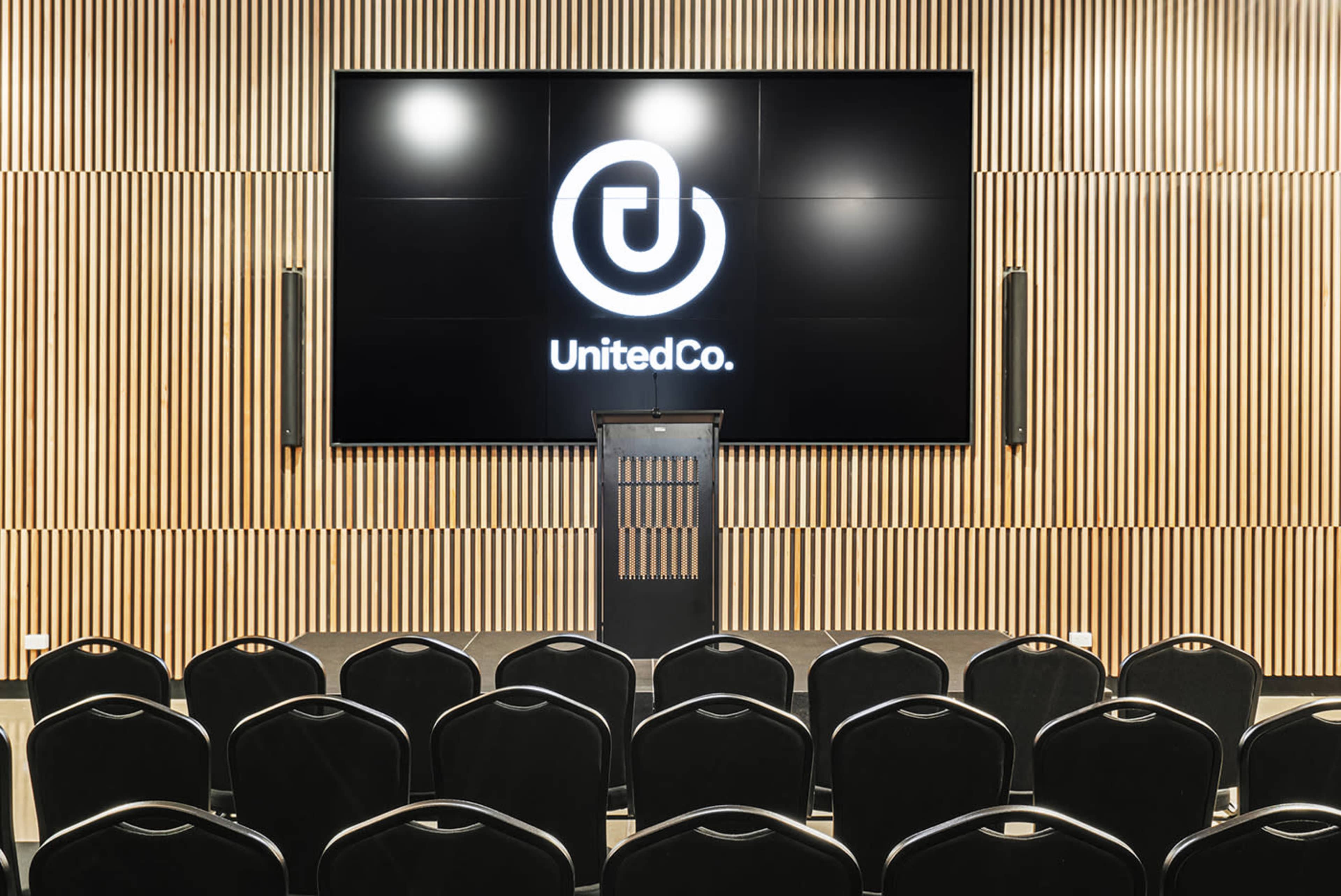 The image shows a conference room set up with black chairs facing a large screen displaying the UnitedCo logo and a podium.