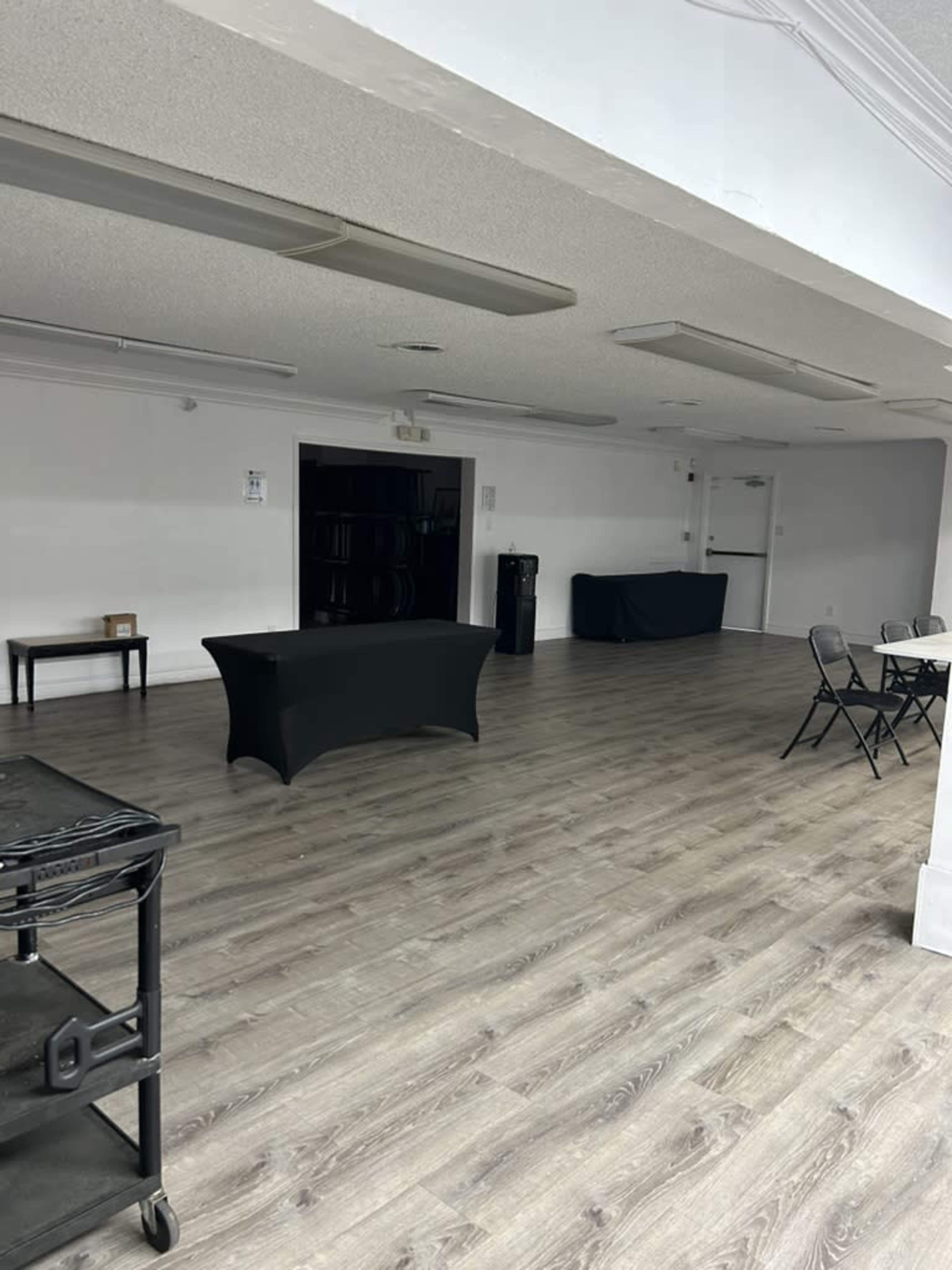 The image shows a large, empty room with a wooden floor, a black table in the center, and folding chairs arranged near the wall.
