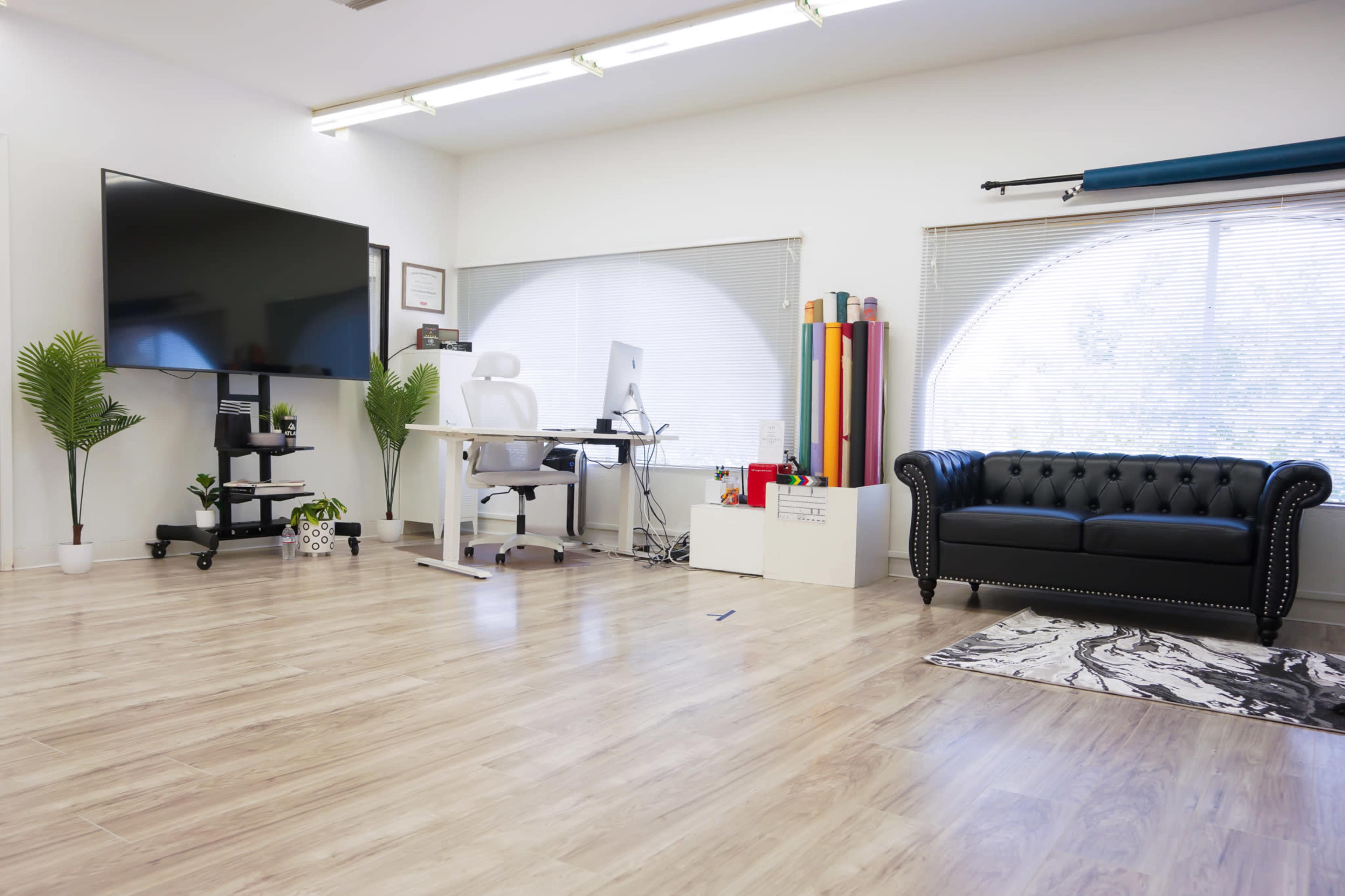 A modern office space featuring a large TV mounted on the wall, a white desk with a computer, a black couch, and various indoor plants.