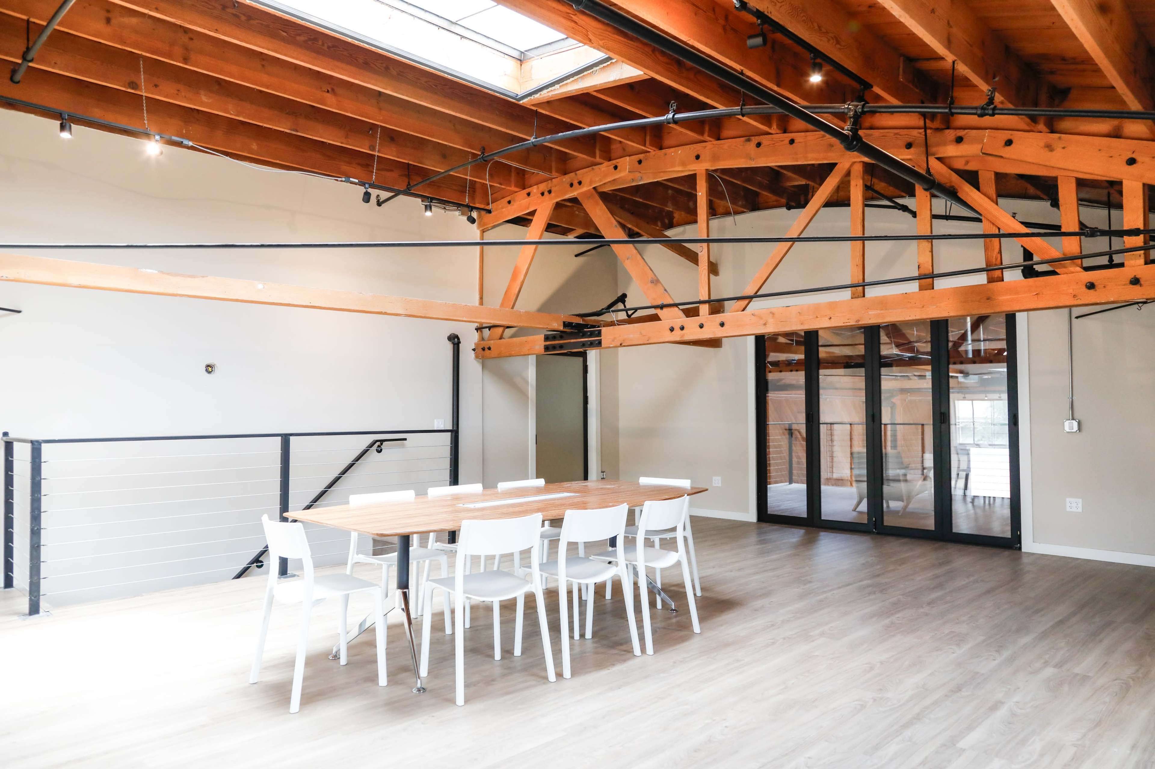 A spacious, modern dining area features a large wooden table surrounded by white chairs, with exposed wooden beams and large windows allowing natural light to flood the space.