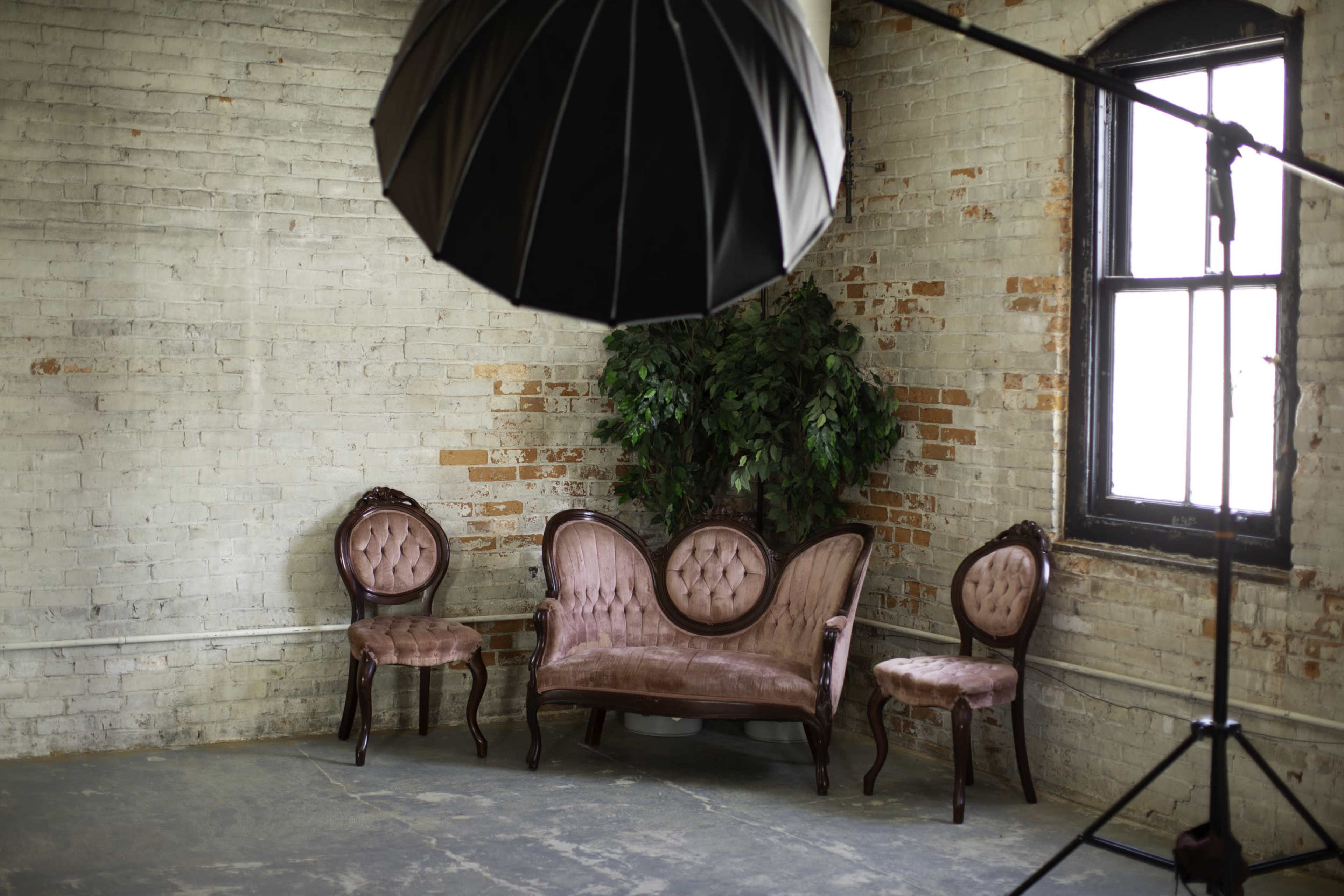 A vintage sofa and three matching chairs are arranged in a corner of a room with exposed brick walls, beneath a large photography light.