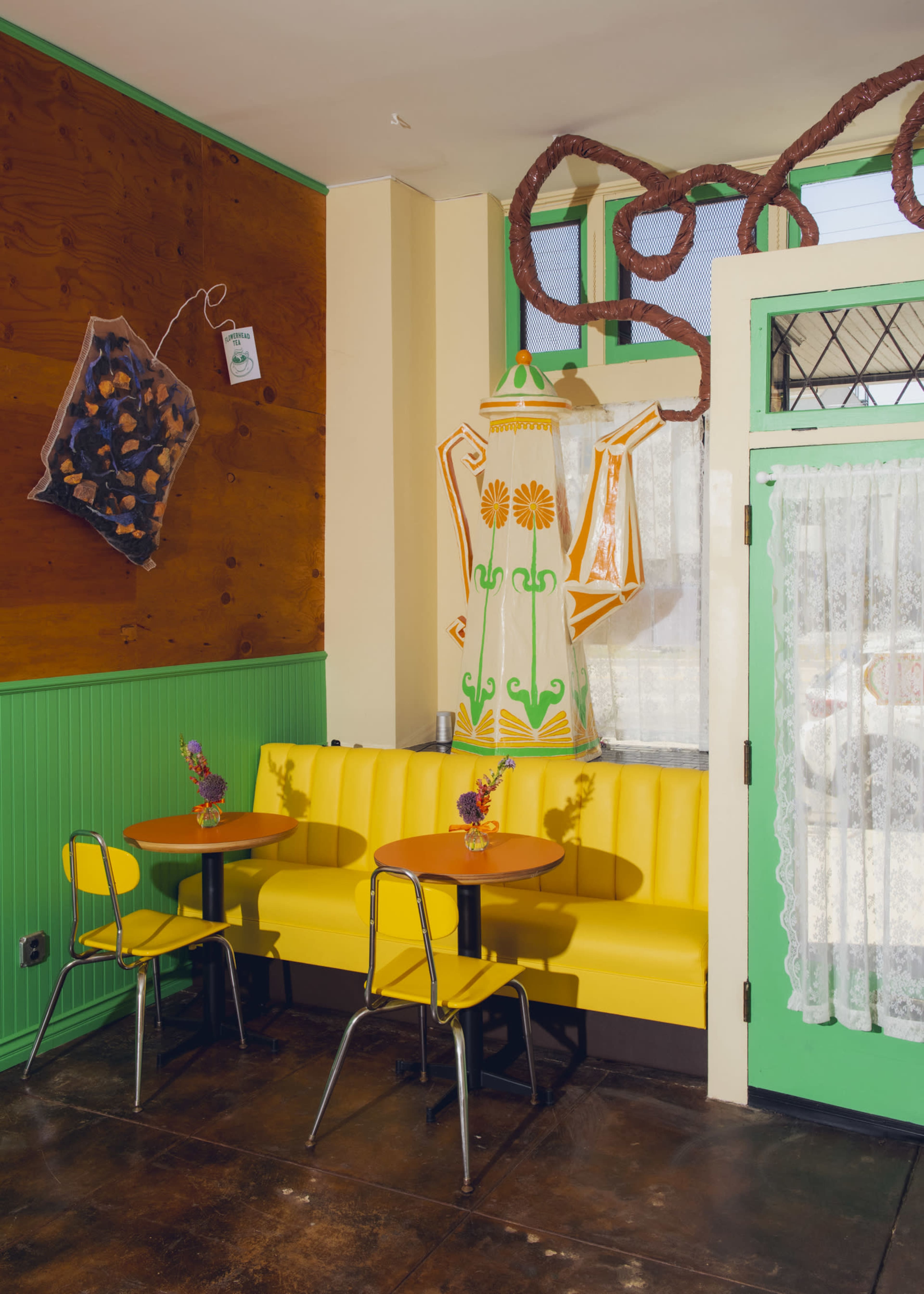 The image shows a brightly colored café corner with a yellow bench, two small black tables, and a large decorative teapot on the wall.