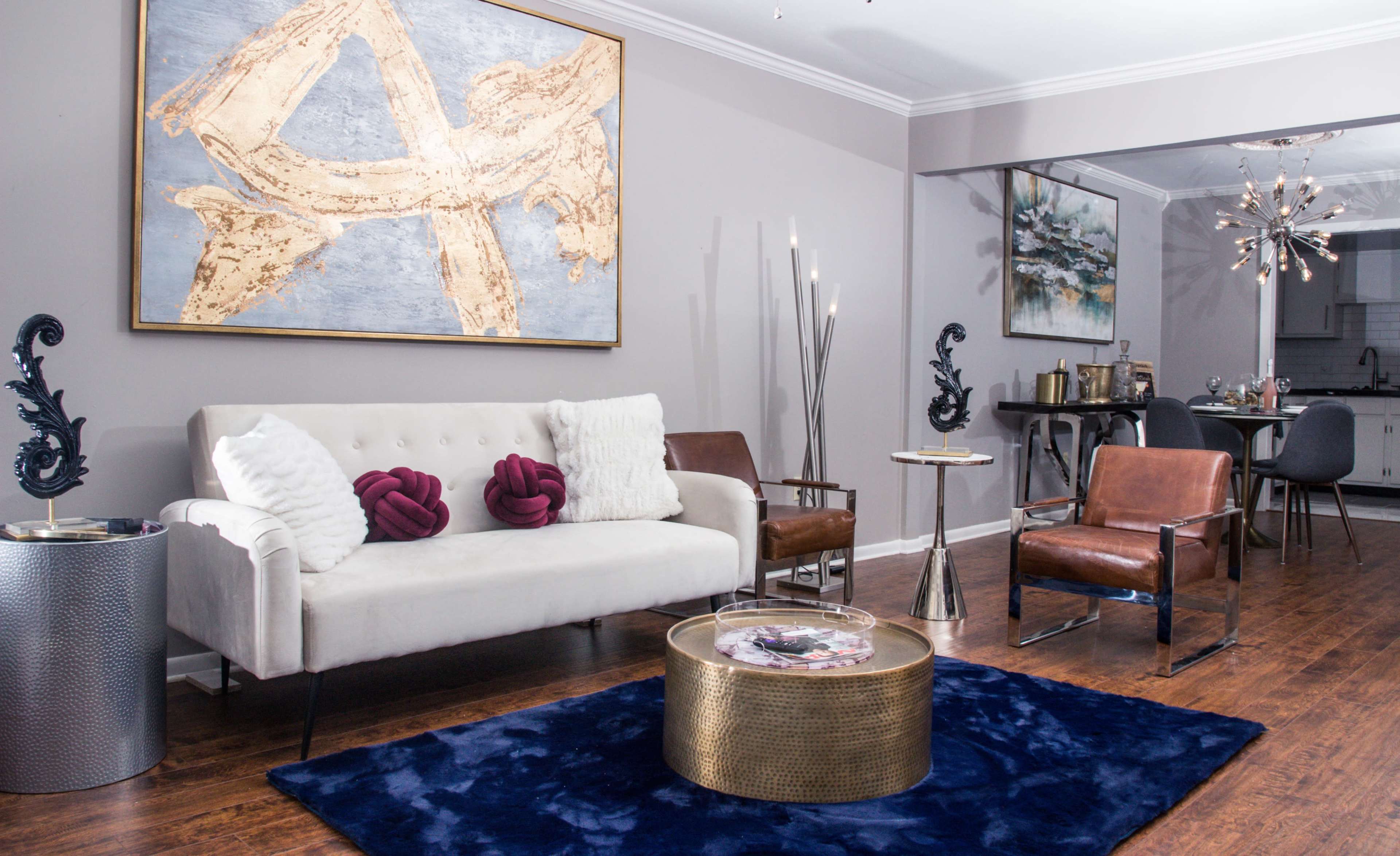A modern living room featuring a white sofa, brown accent chairs, a dark blue rug, and contemporary artwork on the walls.