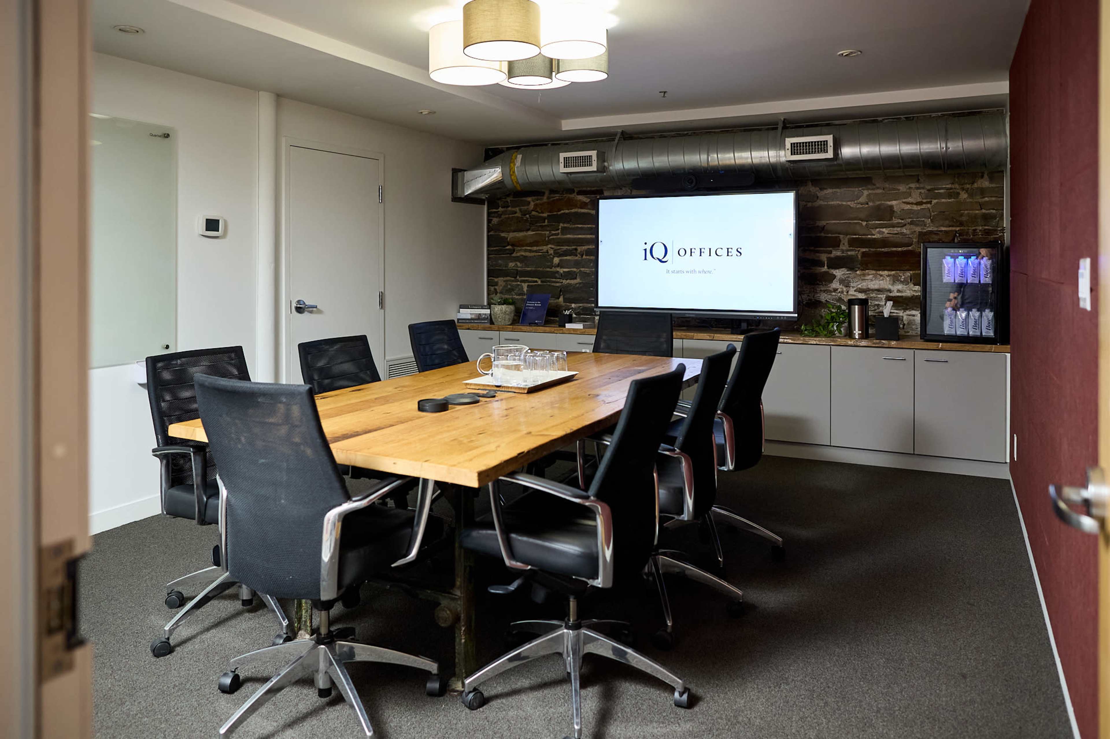 A conference room features a large wooden table surrounded by black swivel chairs, with a display screen on the wall and refreshments in the corner.