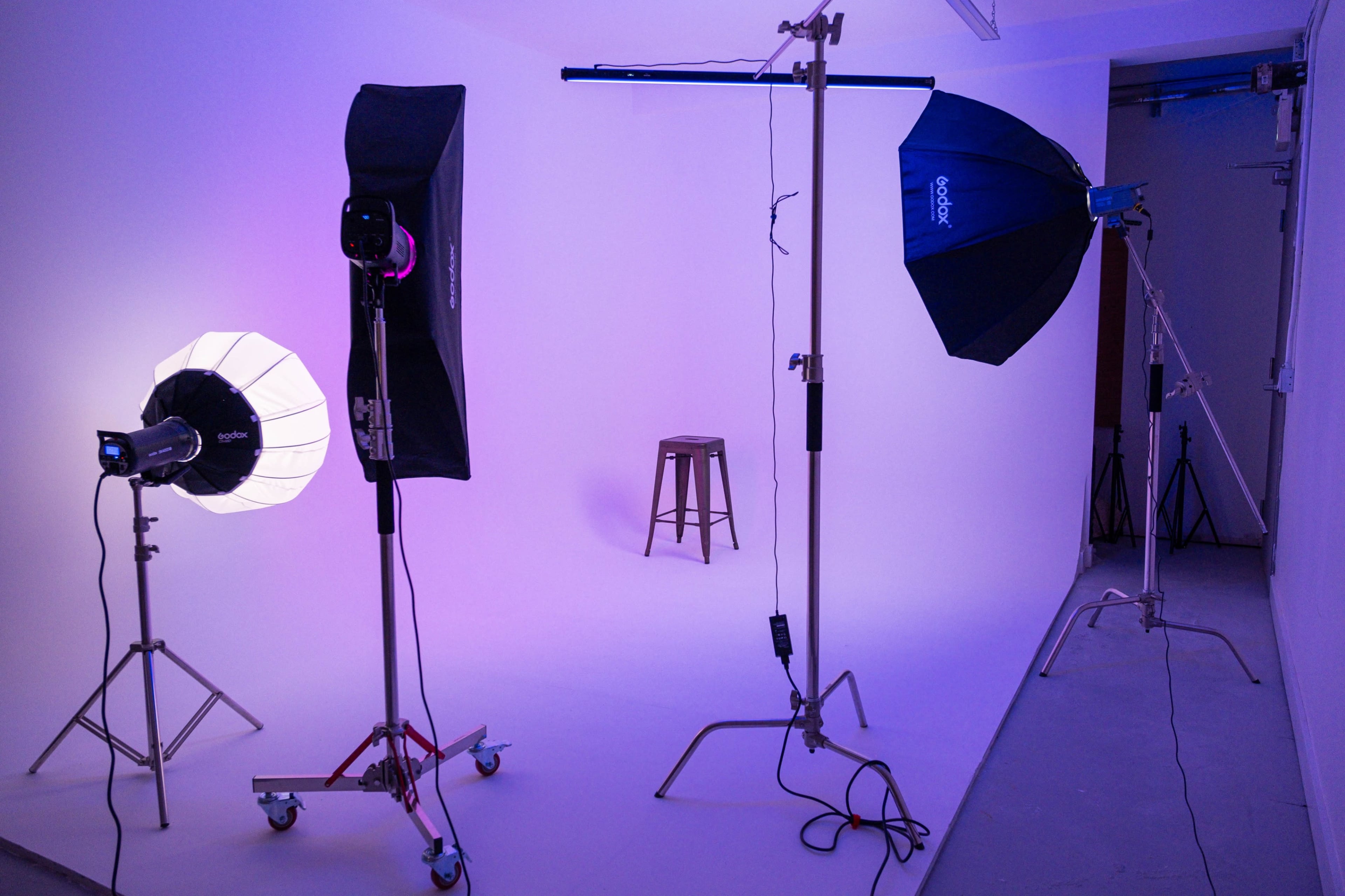 The image shows a photography studio setup with multiple light sources, including a softbox and an umbrella light, and a wooden stool in the center against a white backdrop.