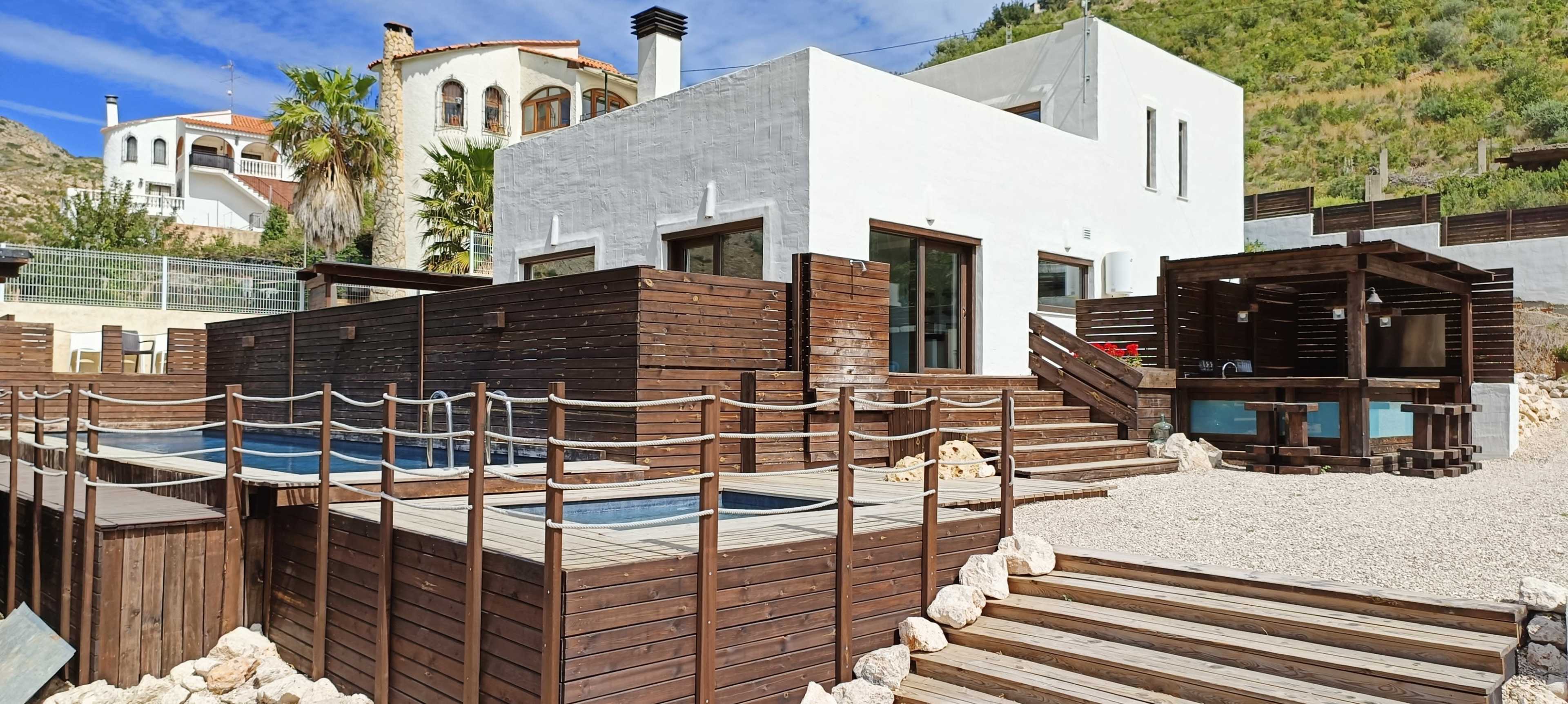 A modern villa with wooden decking and multiple outdoor areas, including swimming pools, set against a hillside.