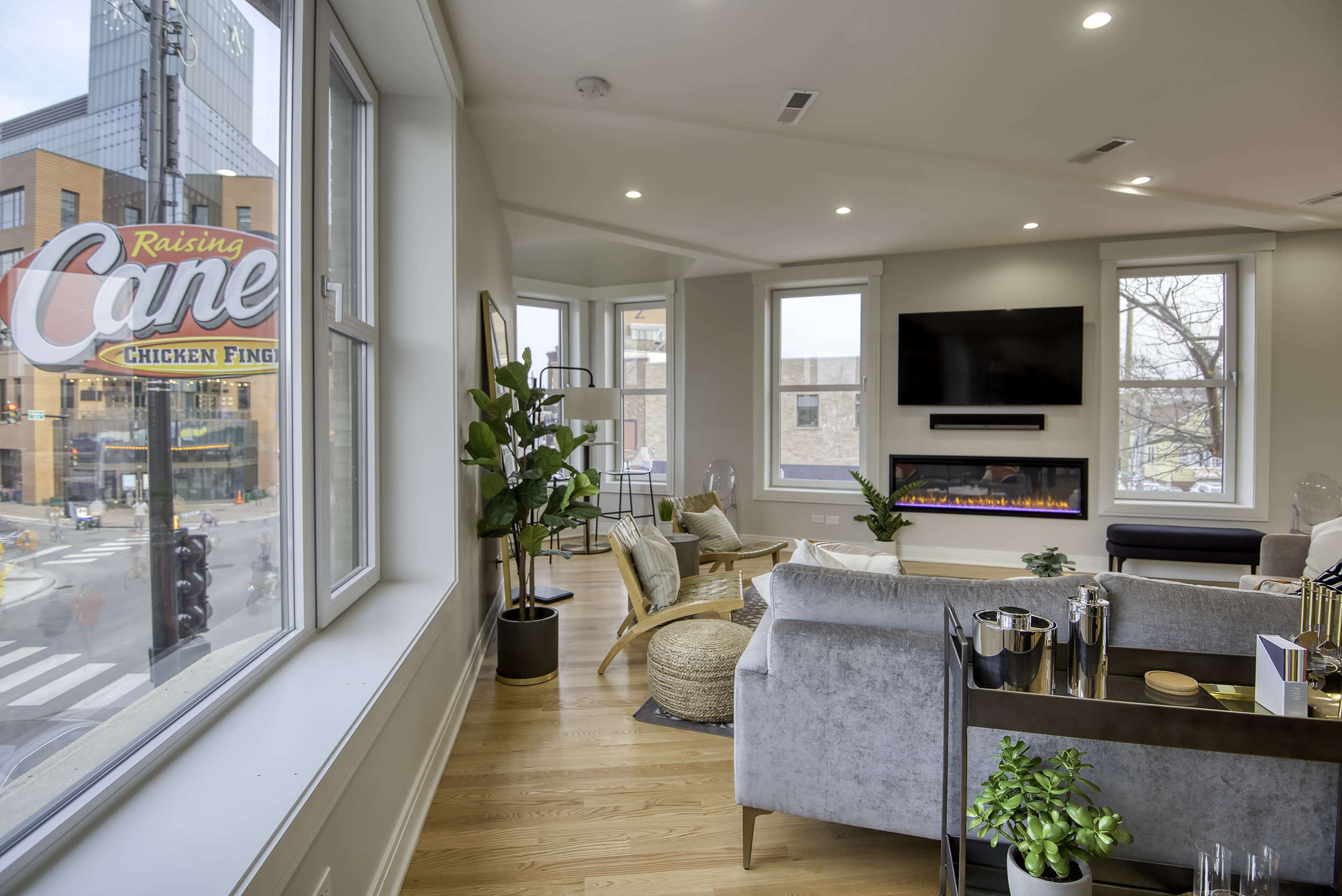 The interior of a modern living room features large windows, contemporary furniture, and a view of a Raising Cane's Chicken Fingers sign outside.