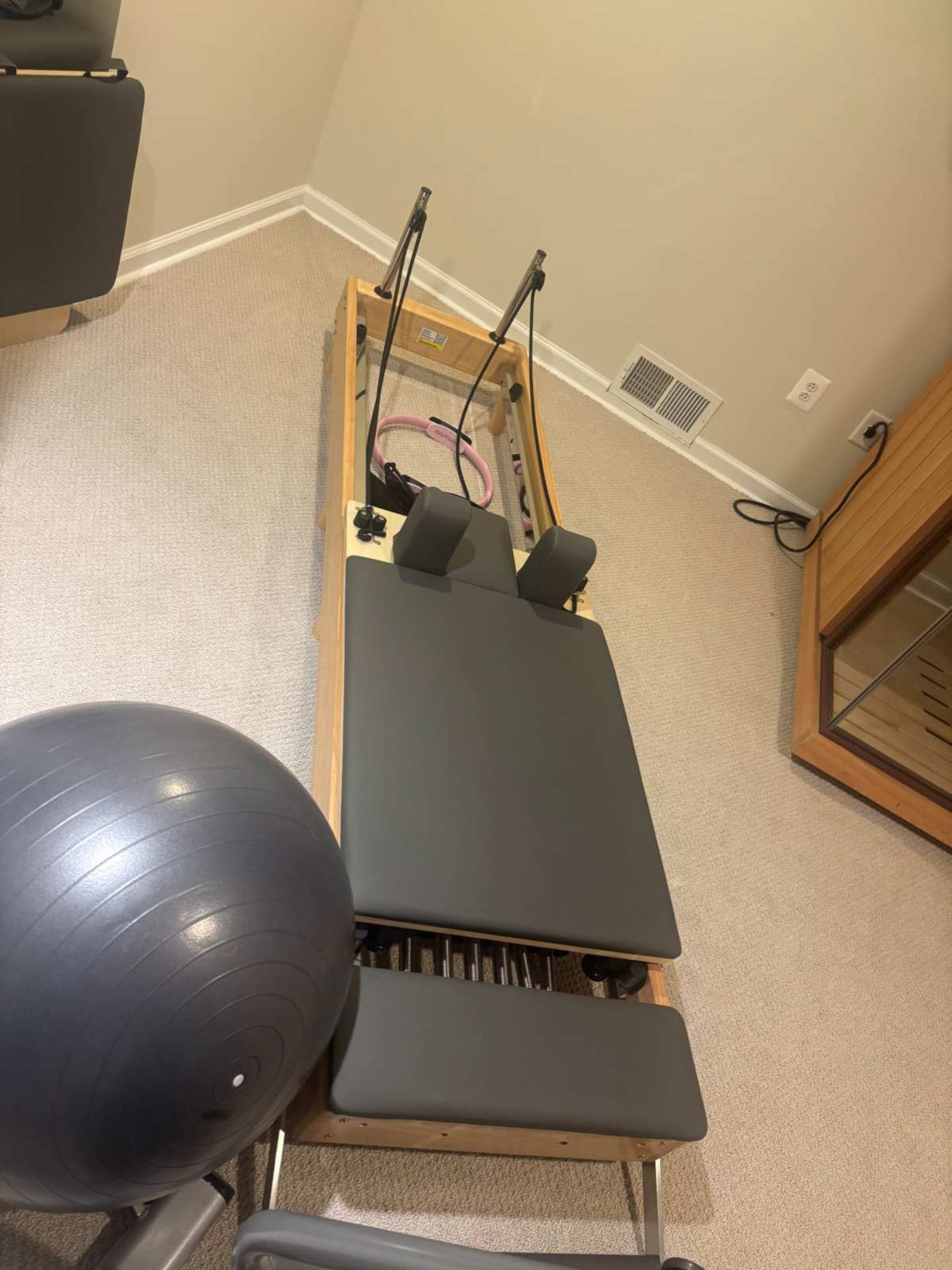 The image shows a wooden Pilates reformer machine with padded components and resistance bands, placed on a carpeted floor near a black exercise ball.