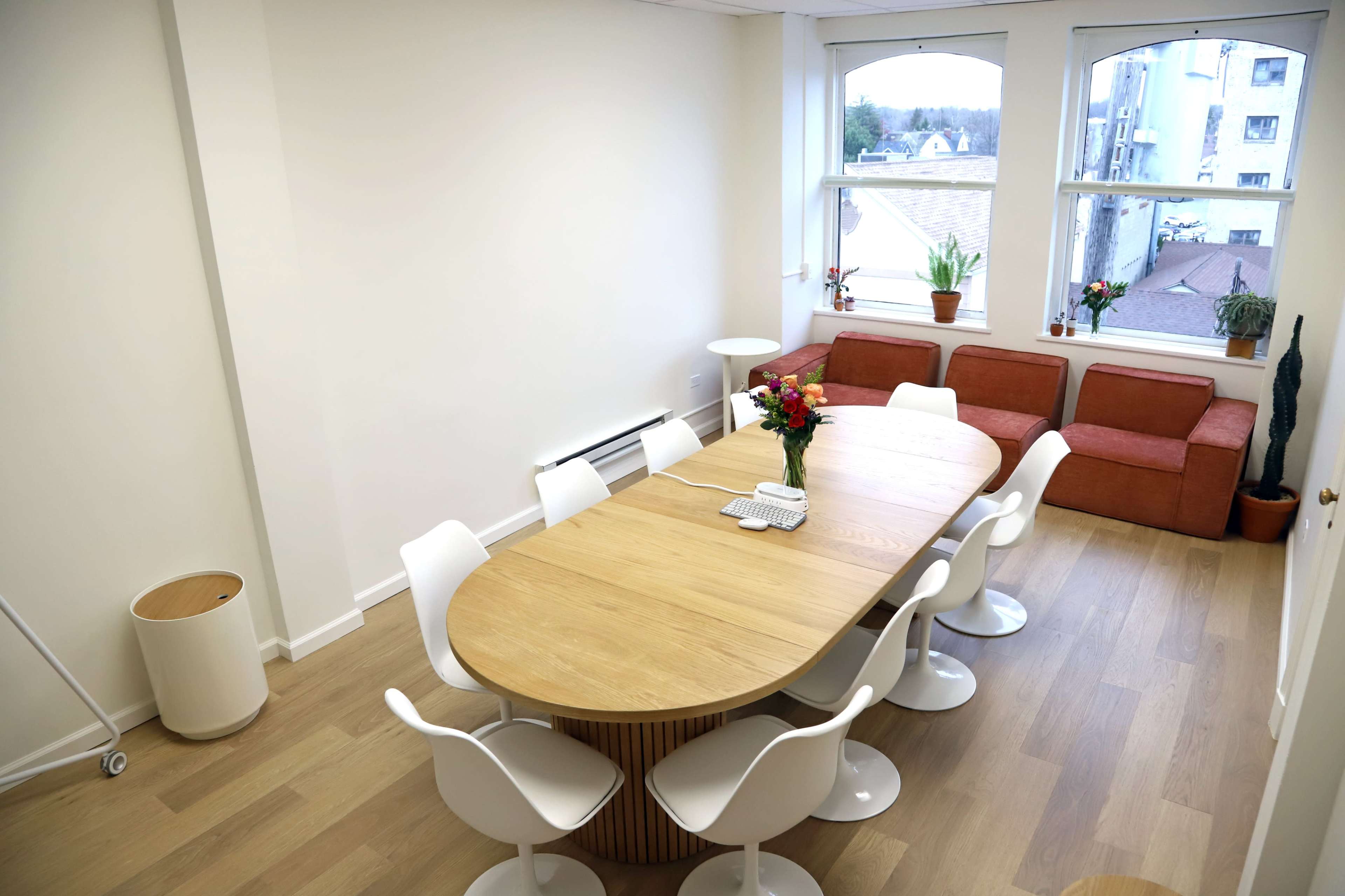 A bright meeting room features a long wooden table surrounded by white chairs, with a cozy seating area and windows letting in natural light.