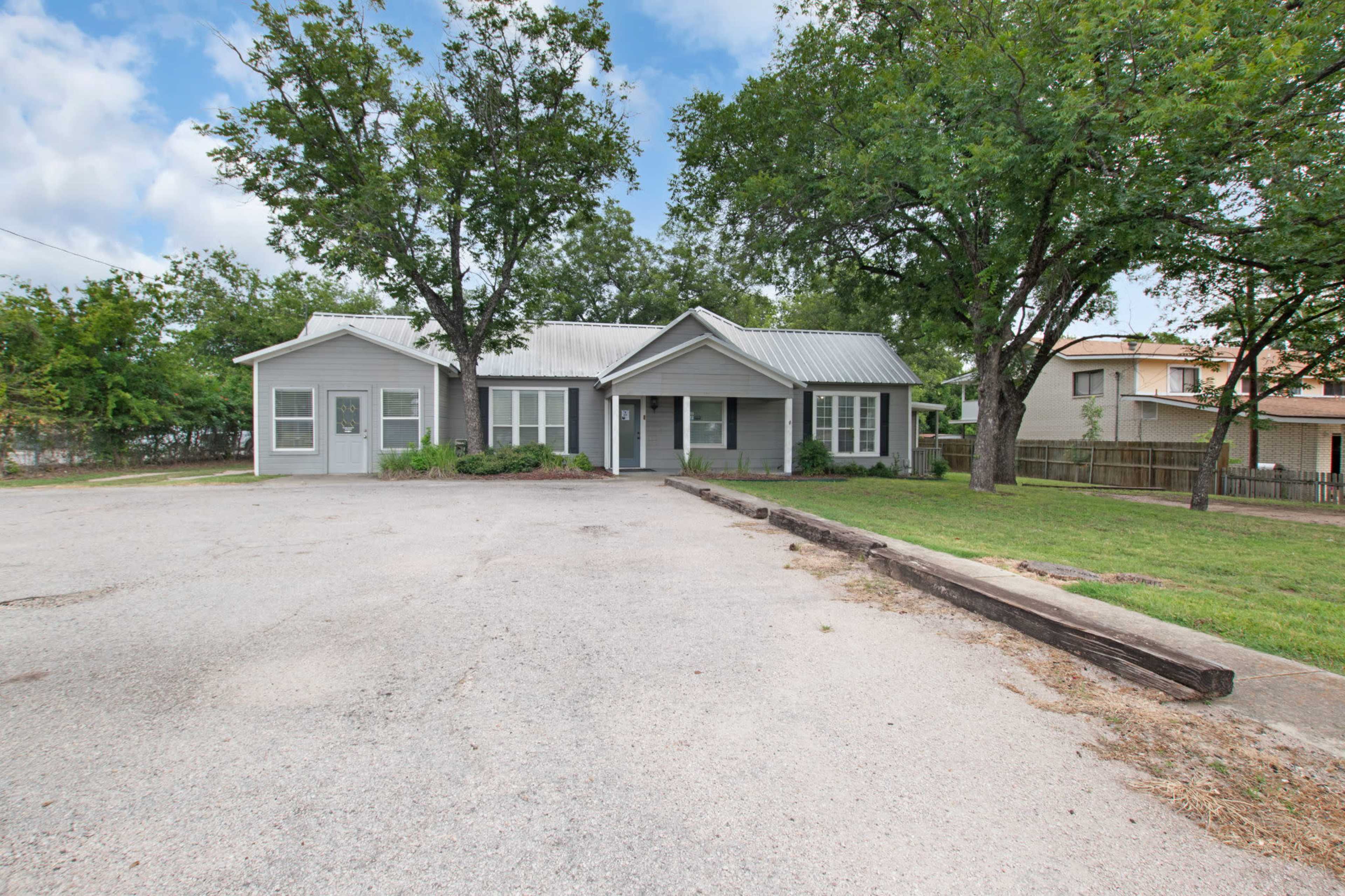 The image shows a single-story house with a metal roof surrounded by trees and a gravel driveway.