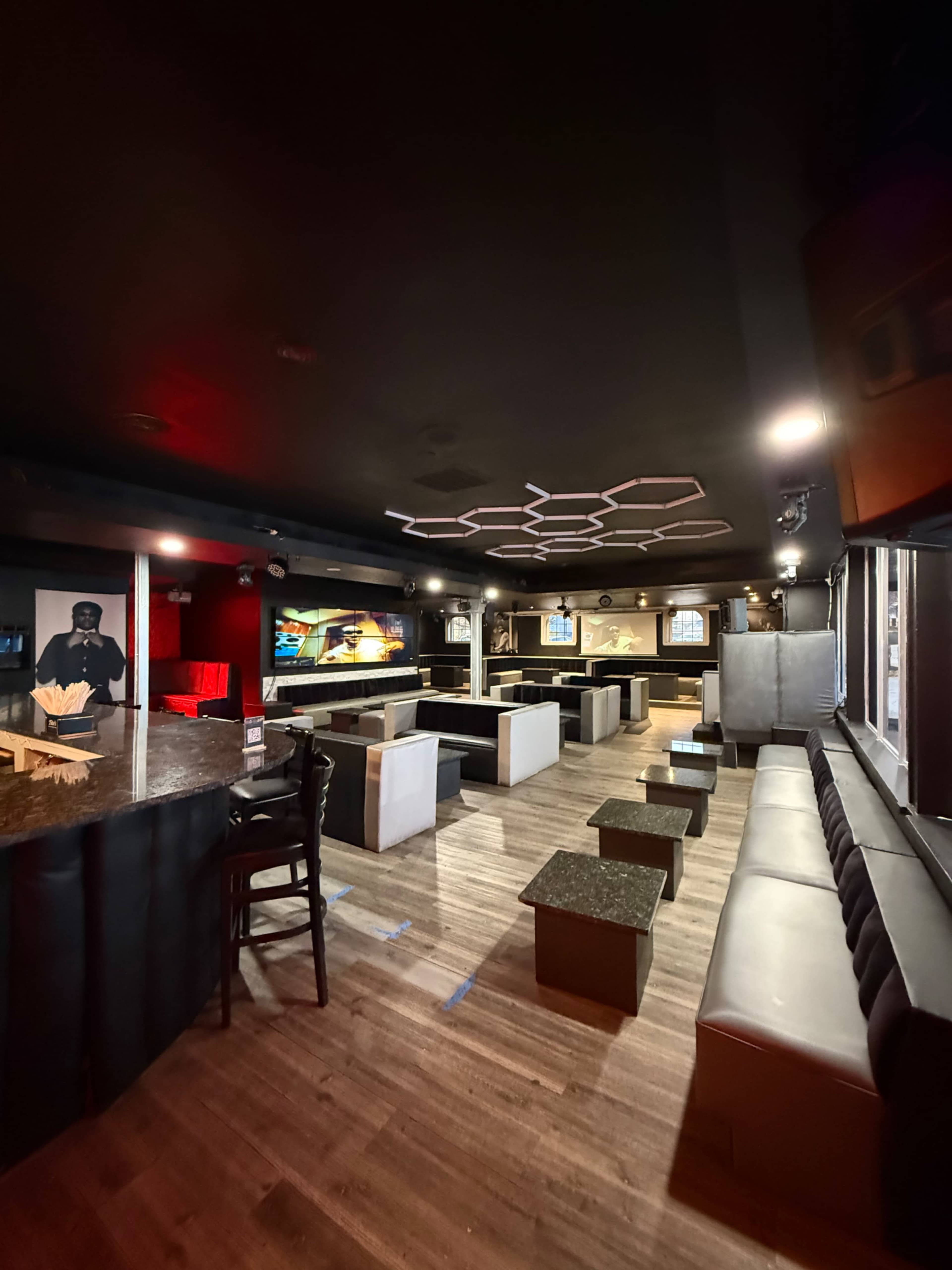 The image shows a dimly lit lounge with rows of black and gray seating, small tables, and a bar area, featuring wall-mounted screens.