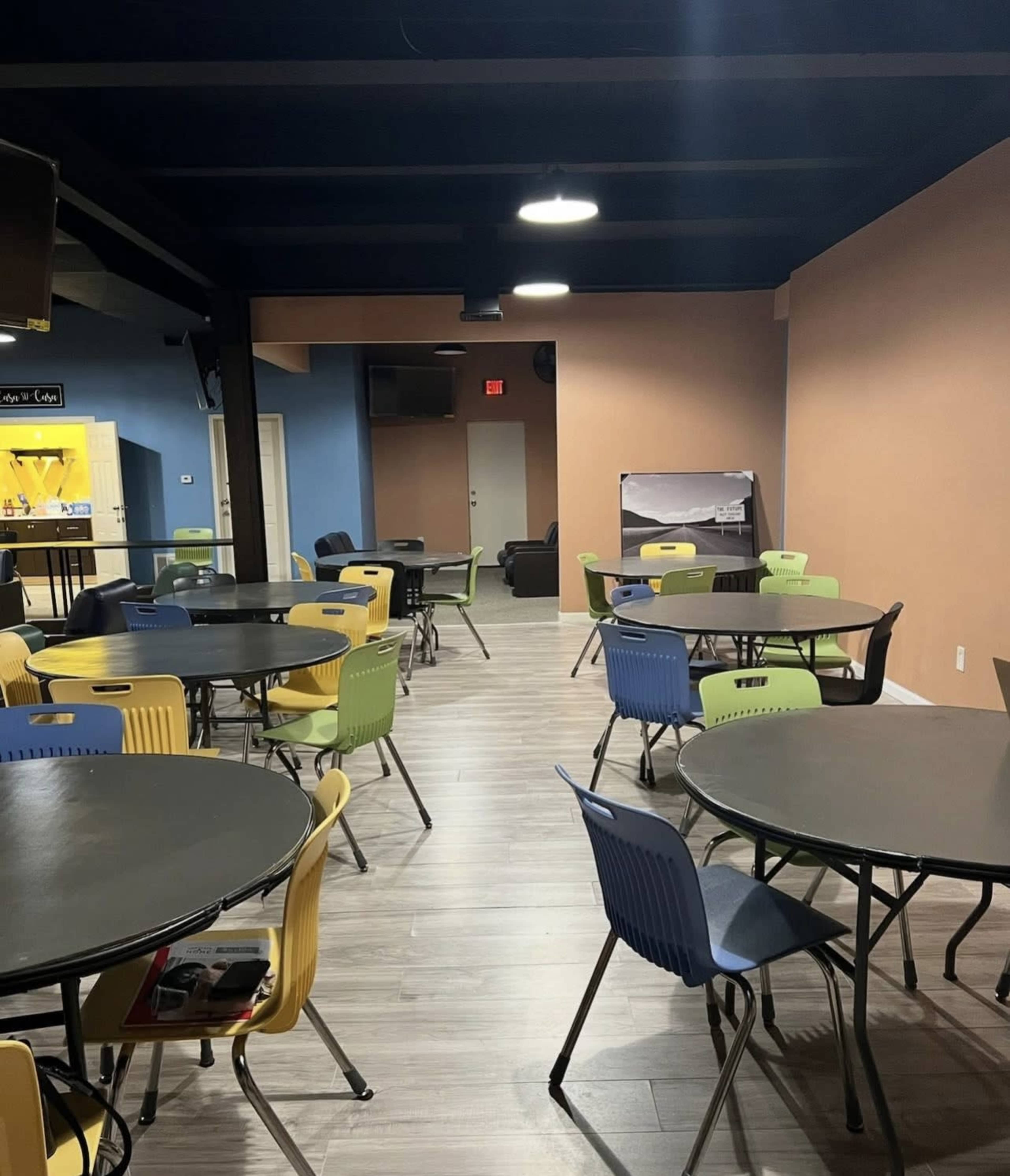 The image shows a brightly lit room with multiple round tables and chairs in various colors arranged throughout the space.