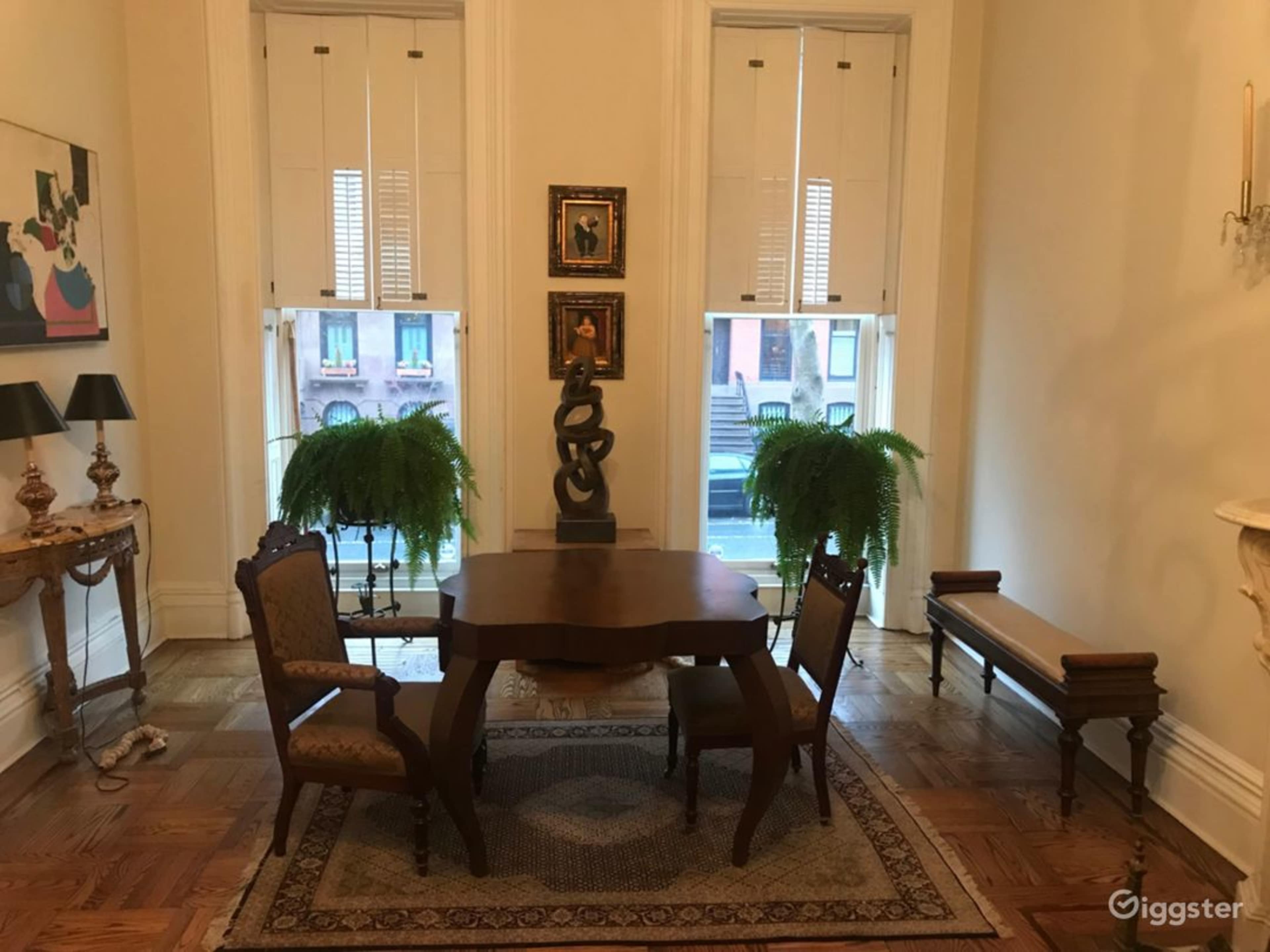 A small, elegantly furnished room features a wooden dining table with four chairs, flanked by two large potted ferns and notable artwork on the walls.