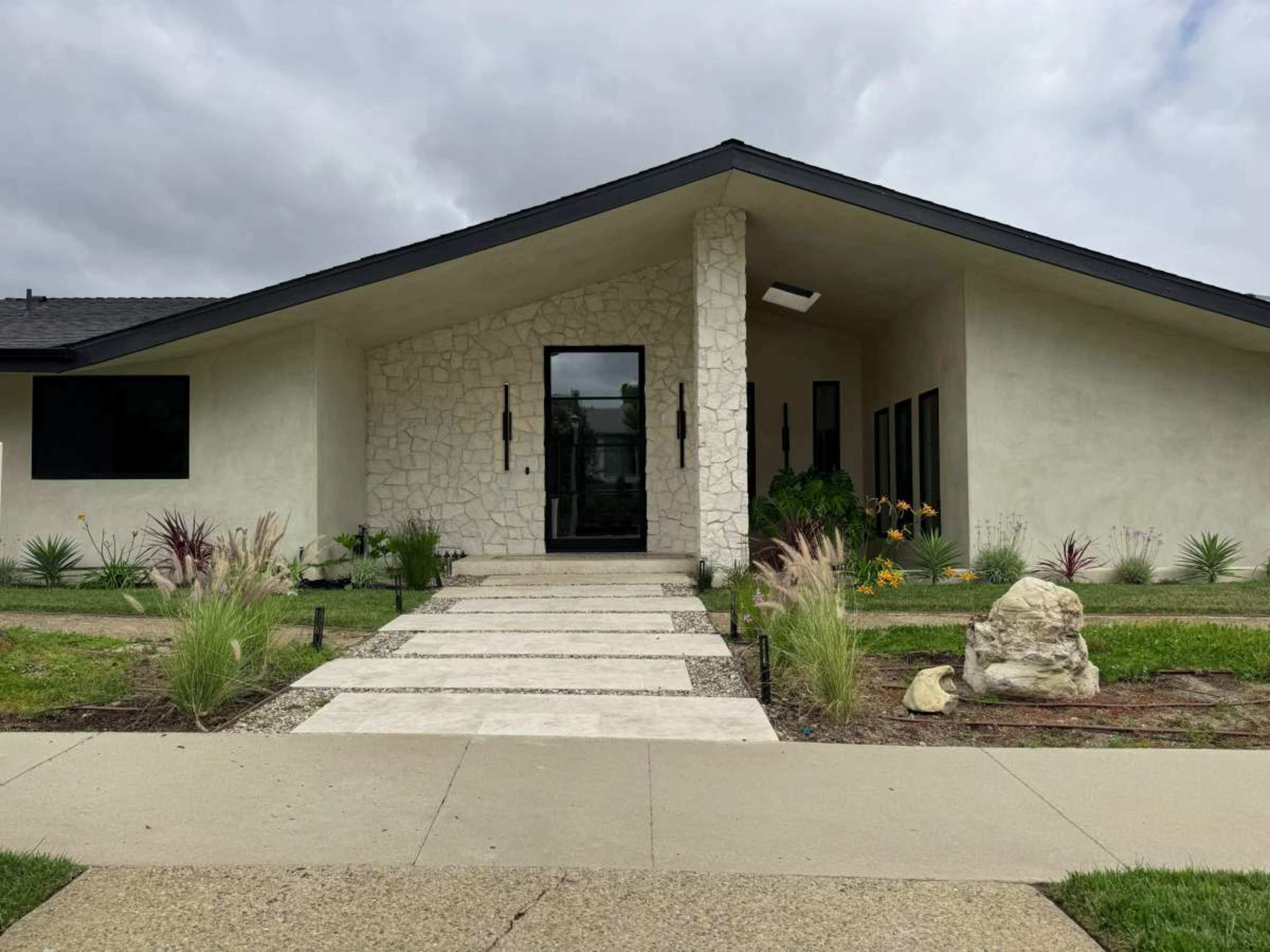 The image shows a modern house with a large entrance, featuring a stone facade, a prominent front door, and landscaped gardens on either side of a concrete pathway.