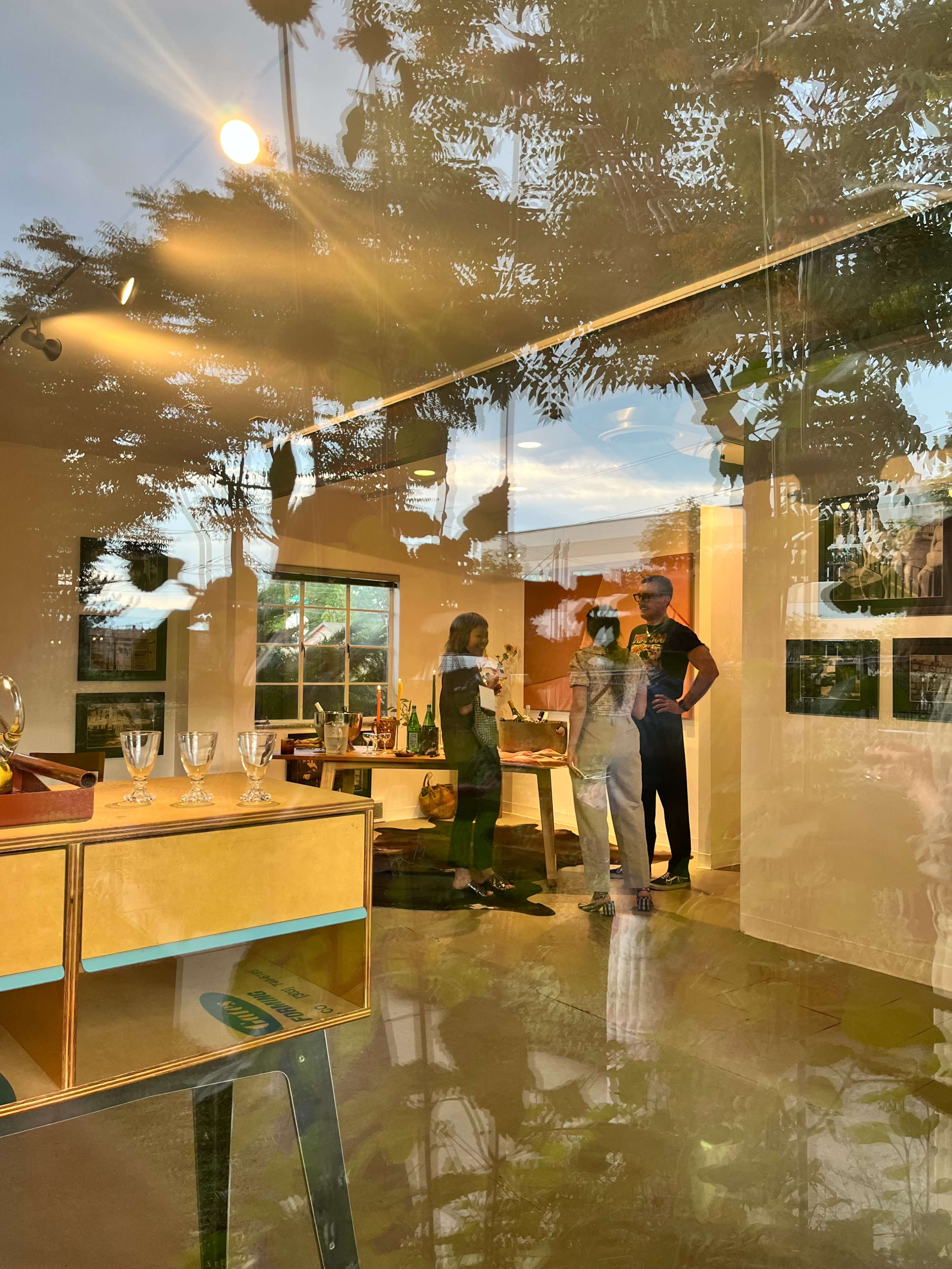 Two people engage in conversation inside a bright, modern room furnished with a table and glassware, while reflected greenery outside creates a layered visual effect on the glass window.