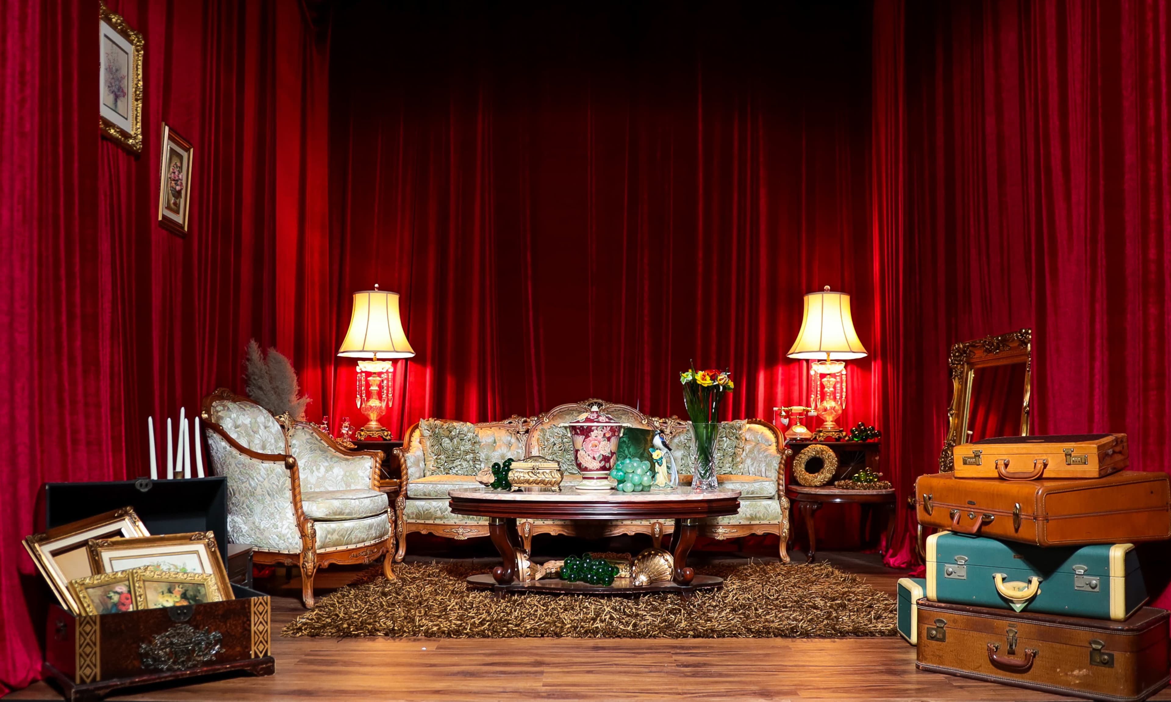 The scene features a vintage-style living room with a red curtain backdrop, decorated with ornate furniture, lamps, a central table, and various pieces of luggage.