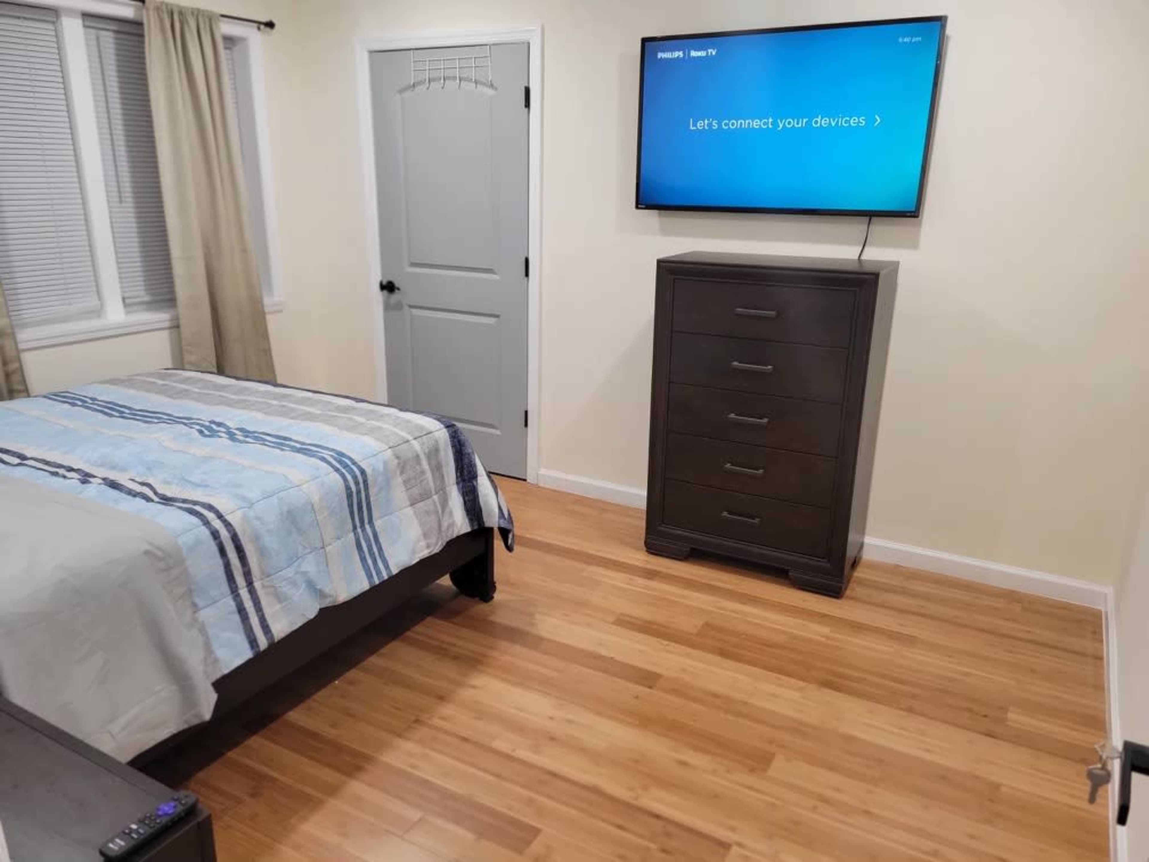 The image shows a bedroom with a wooden bed, a television mounted on the wall, and a dresser, all set against a light-colored wall.