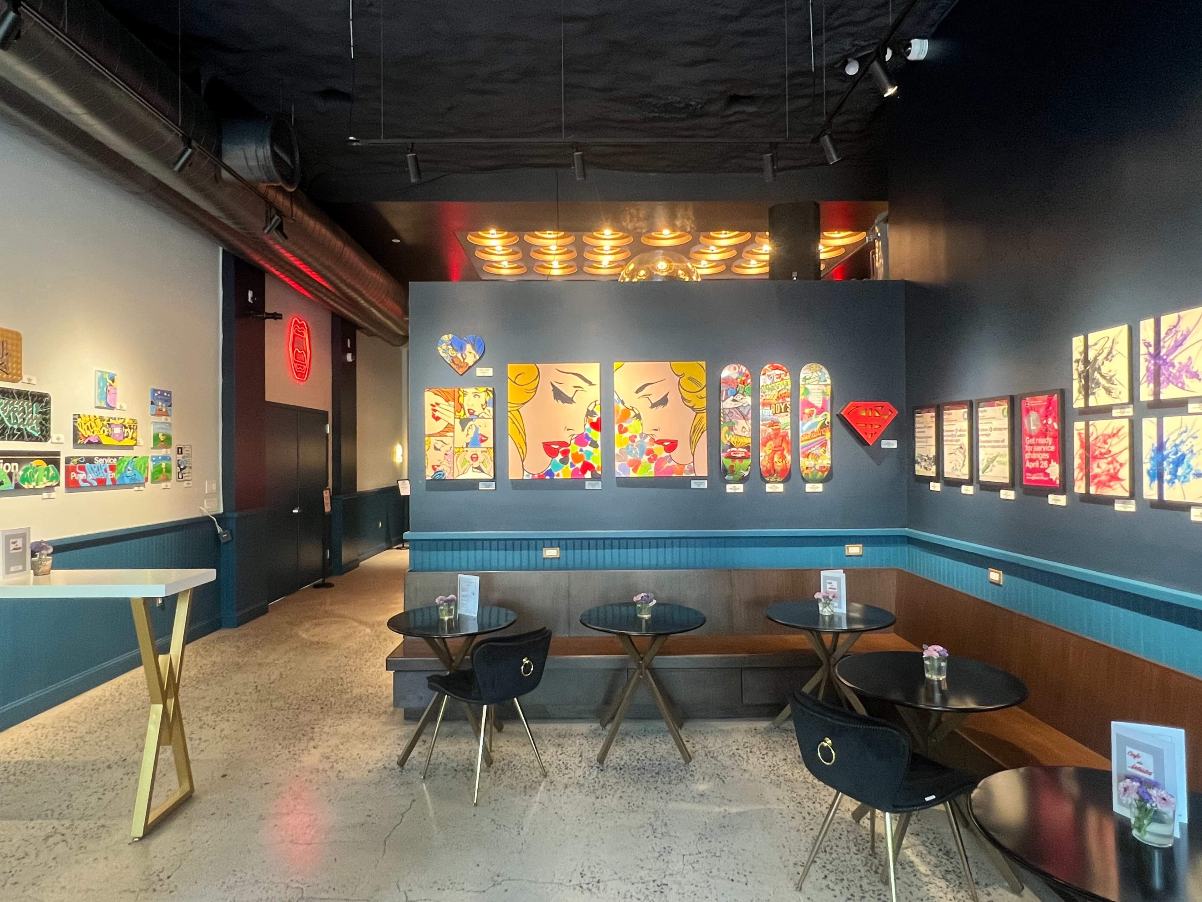 The image shows a modern cafe with a vibrant decor, featuring colorful artwork on the walls and several small tables arranged for seating.