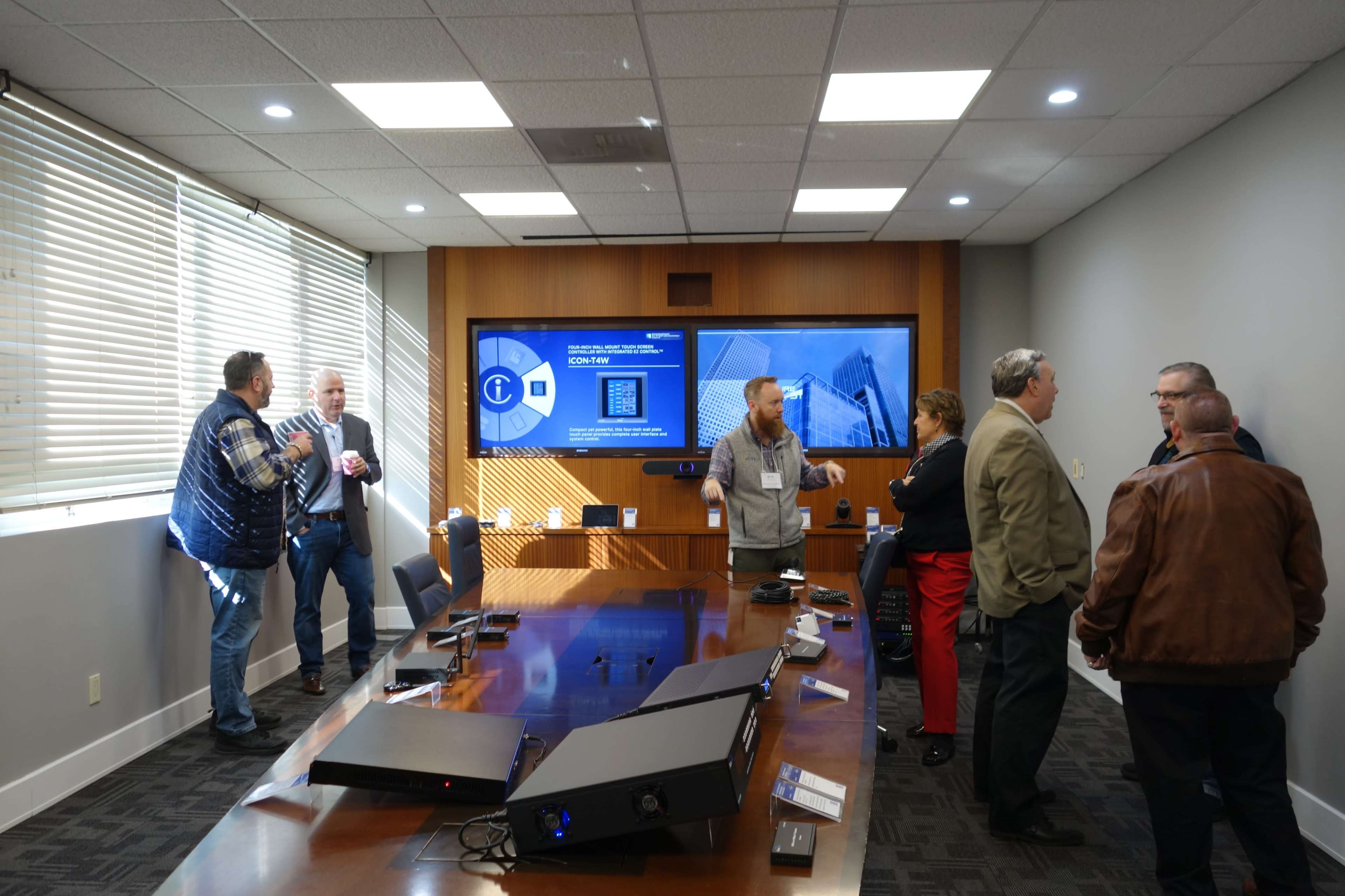 A group of professionals is engaged in conversation around a large conference table in a well-lit meeting room featuring a digital display.