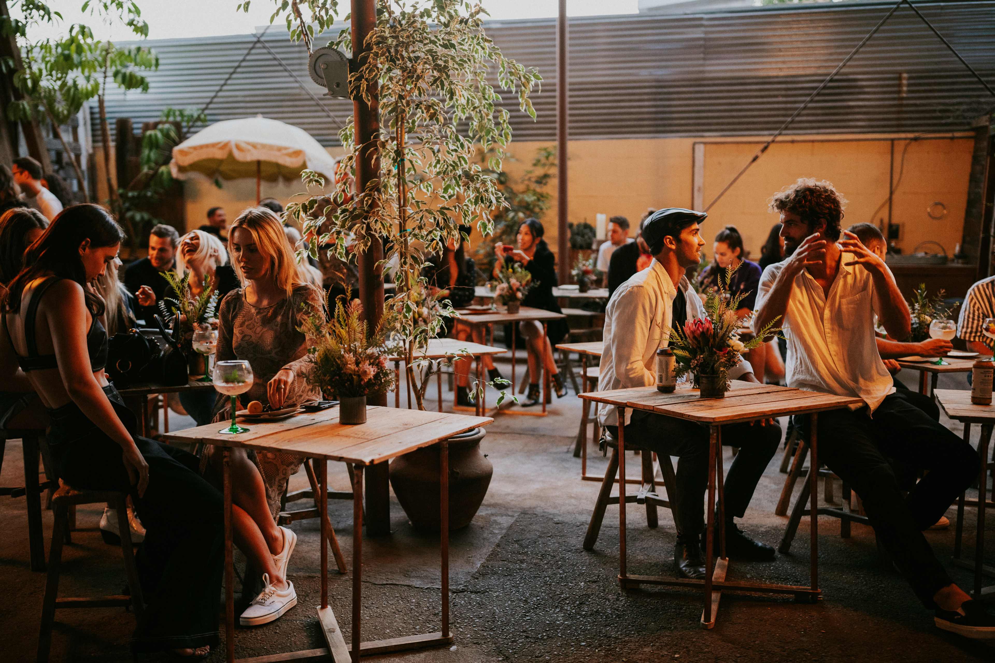 A lively gathering in an outdoor café with people seated at wooden tables surrounded by plants.
