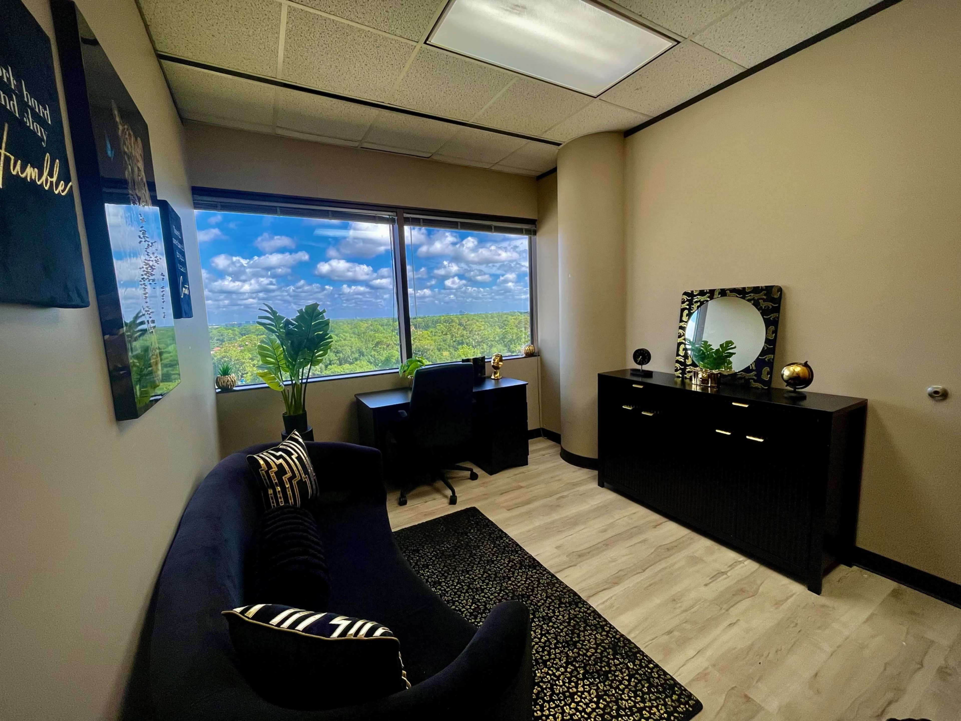 The image shows an office space with a dark couch, a desk and chair, a dresser, and large windows overlooking a landscape with clouds.
