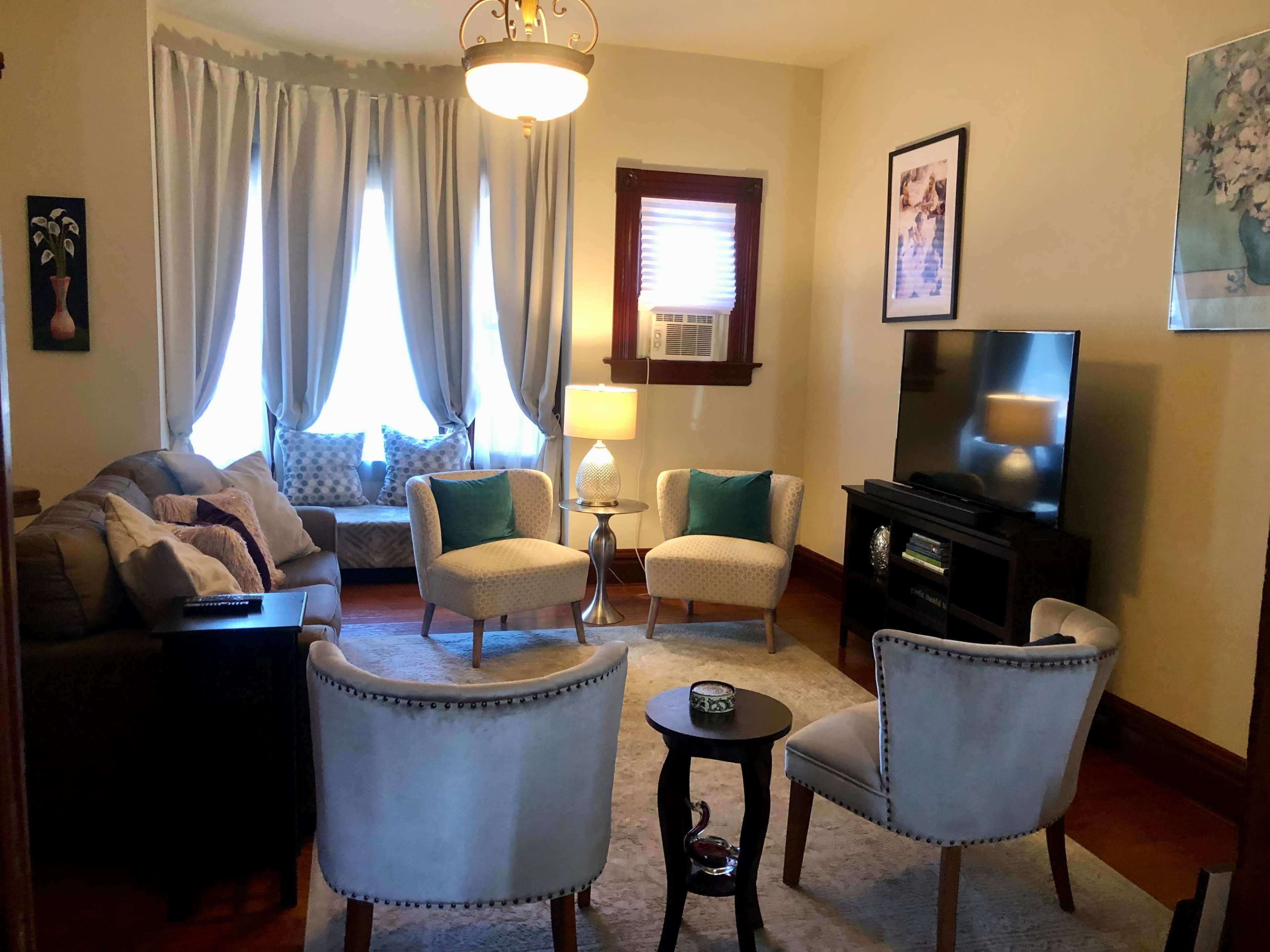 A cozy living room with two armchairs and a sofa arranged around a small table, a television against the wall, and large windows dressed with light curtains.