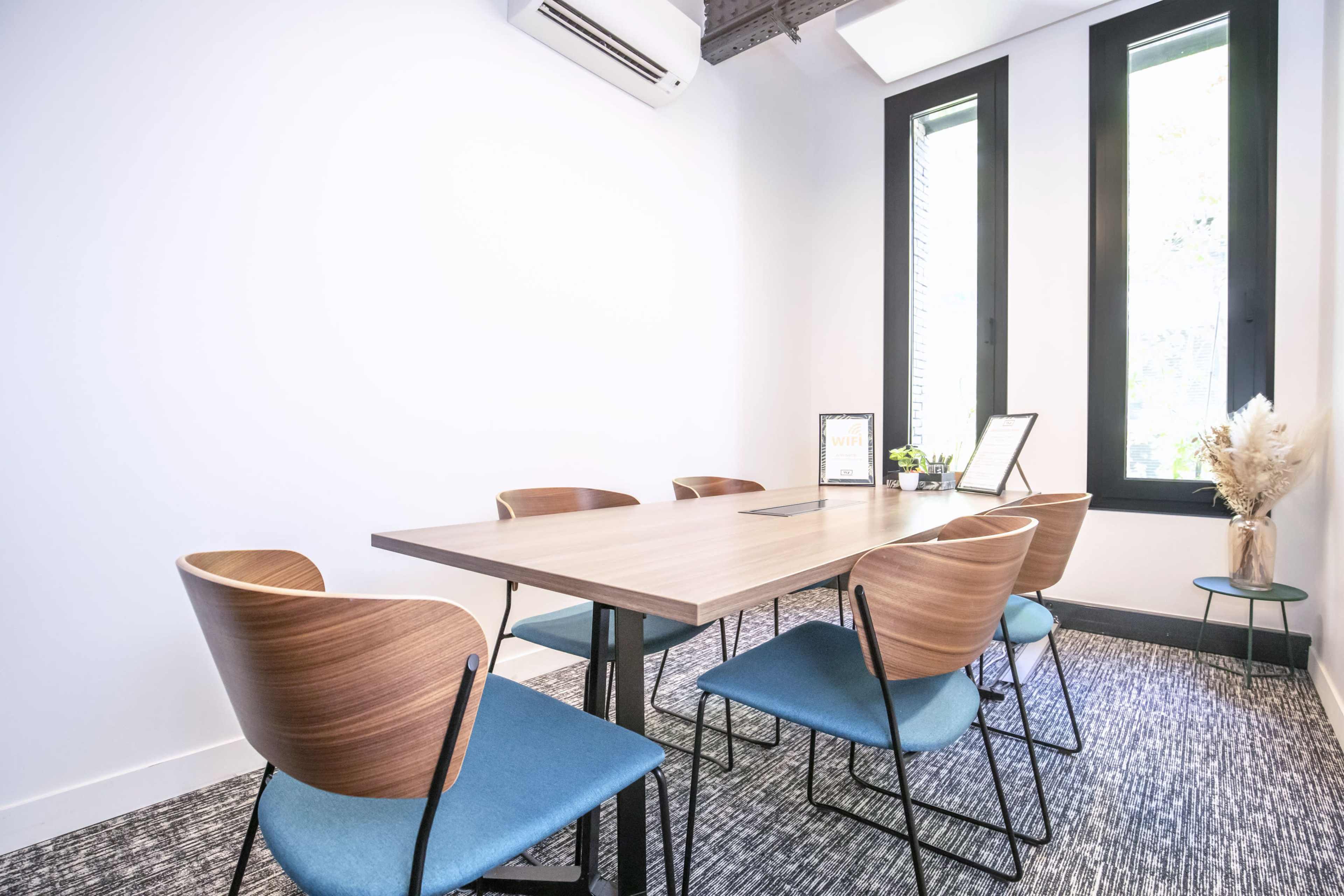 A modern conference room features a wooden table surrounded by six chairs, with large windows allowing natural light in.
