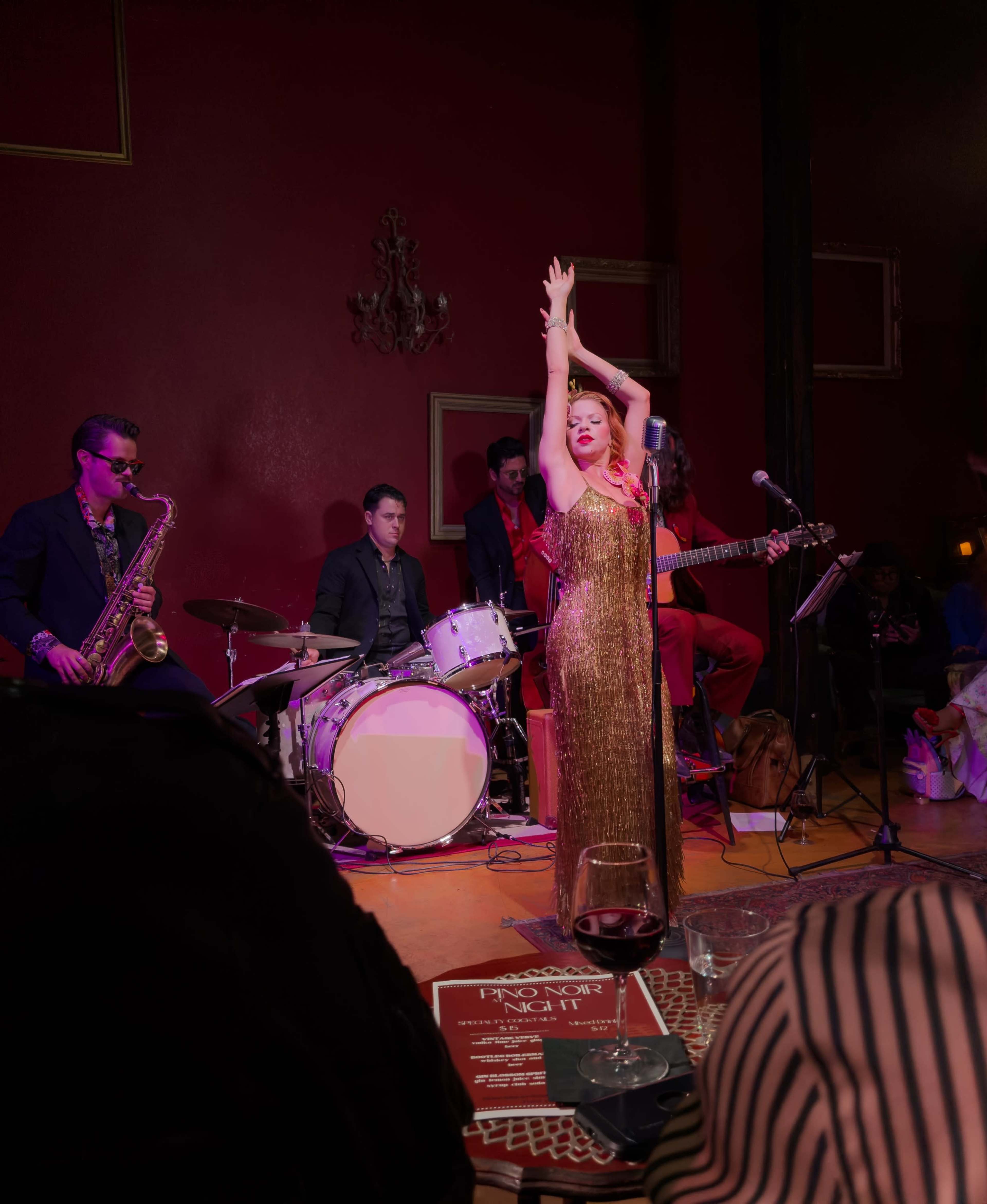 A singer in a glittering dress performs on stage with a jazz band, while an audience enjoys drinks at tables in the foreground.