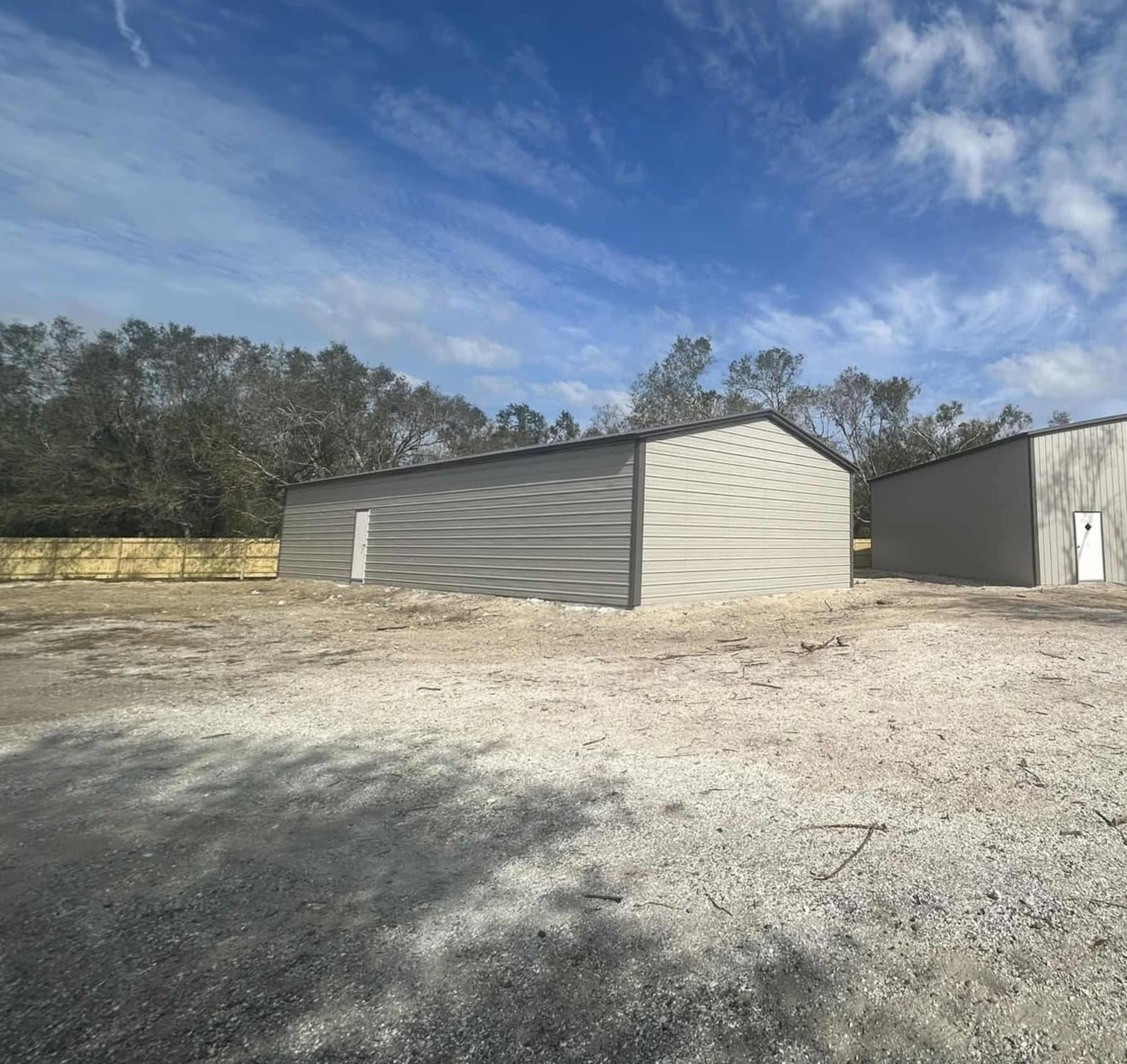 A gray metal building sits on a gravel area, surrounded by trees and blue sky.
